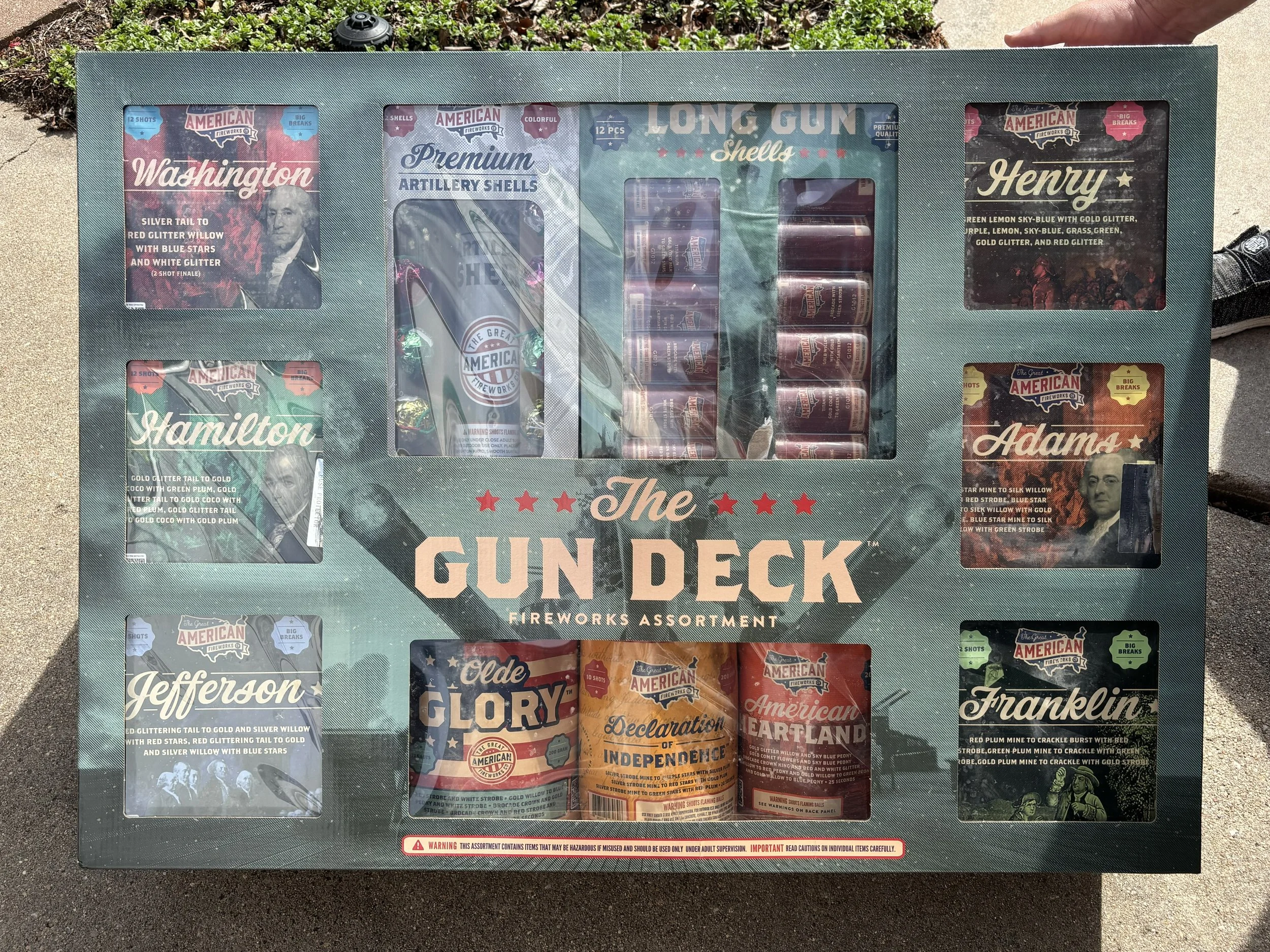 A boxed set of fireworks labeled 'The Gun Deck' with various smaller boxes depicting different fireworks brands and types, including images of people, with a warning label at the bottom.