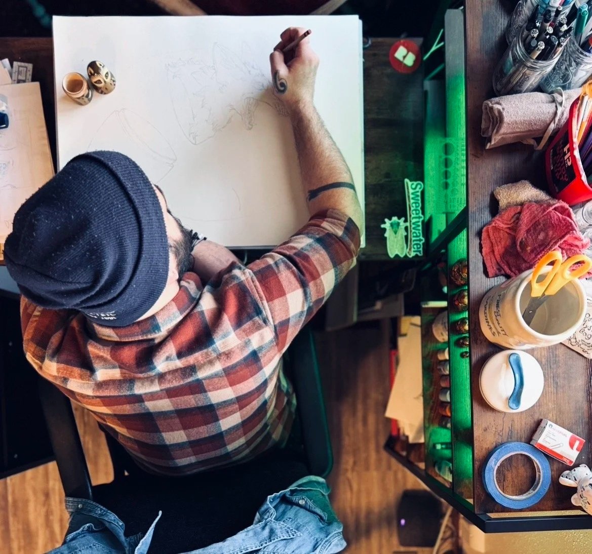 Top-down view of Dale Krause drawing on a large sheet of paper at a cluttered wooden desk, wearing a black beanie and plaid shirt, with art supplies around.