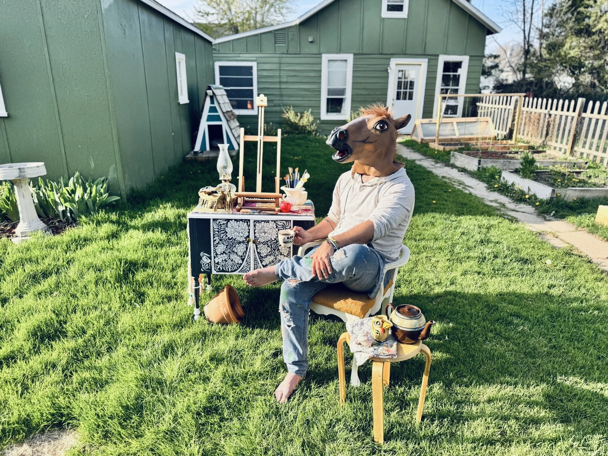 A person sitting outdoors on a white chair, wearing a horse mask, with a table in front of them holding art supplies and a teapot. There is a second small table with a teapot and a decorative item near their feet, in a backyard with grass, a green house, and garden beds.