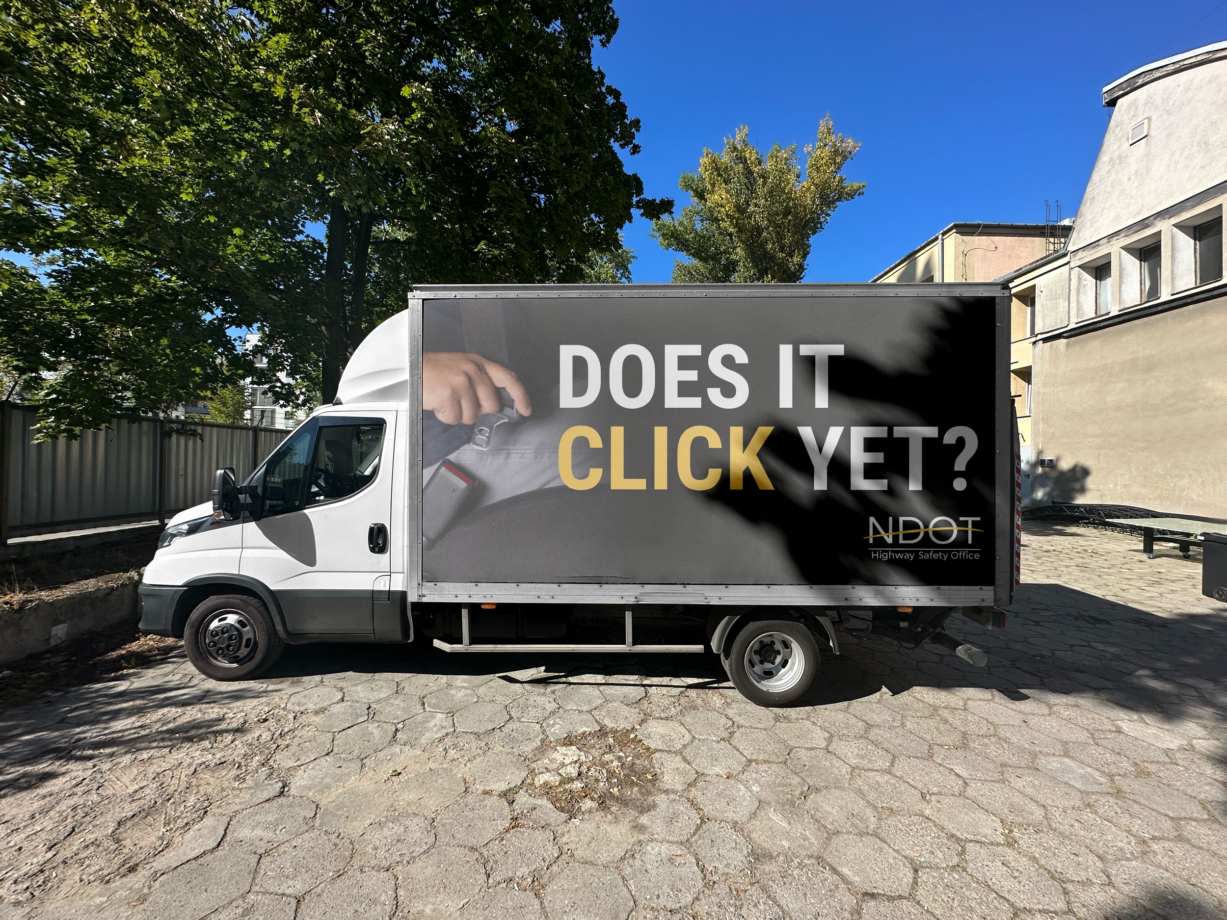 A Nebraska Department of Transportation truck advertisement parked on a paved area. The truck has a black sign on the side with white and yellow text asking, 'Does it click yet?' and a graphic of a hand pressing a doorbell.