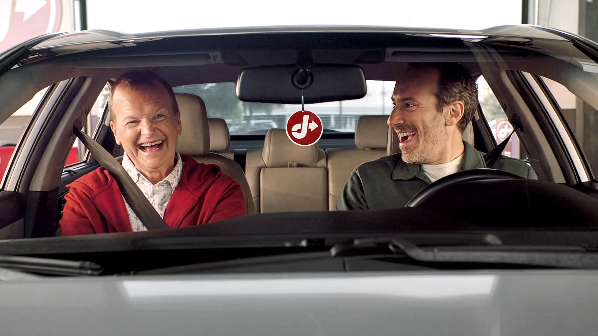 Two advertising icons, previously for Sonic now for Jiffy Lube, sitting in a car, both smiling and laughing, with Peter Grosz in the driver's seat and T.J. Jagodowski in the passenger seat.