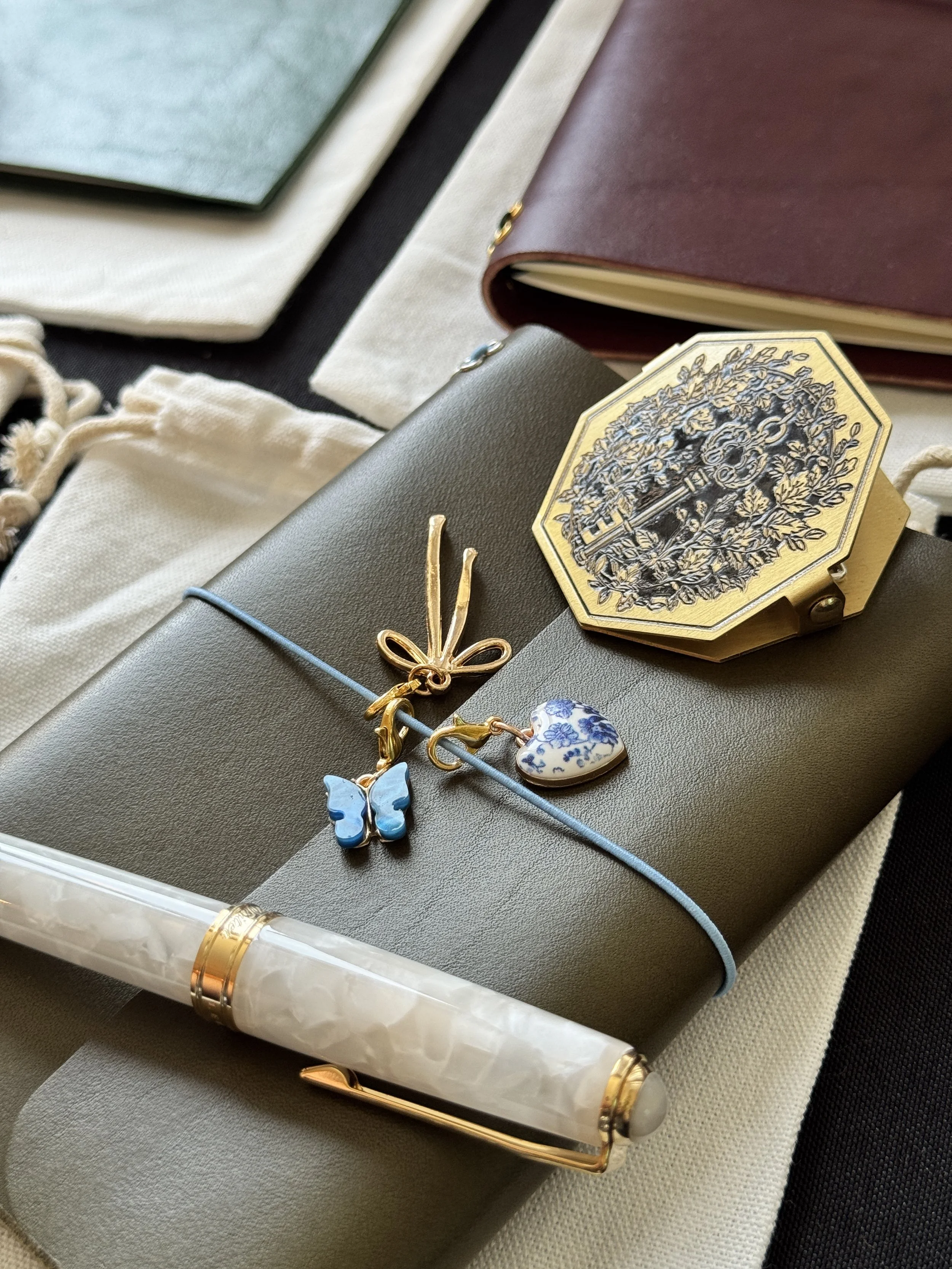 A white fountain pen set against an olive-colored leather journal, with three charms. A brass clip set atop it.