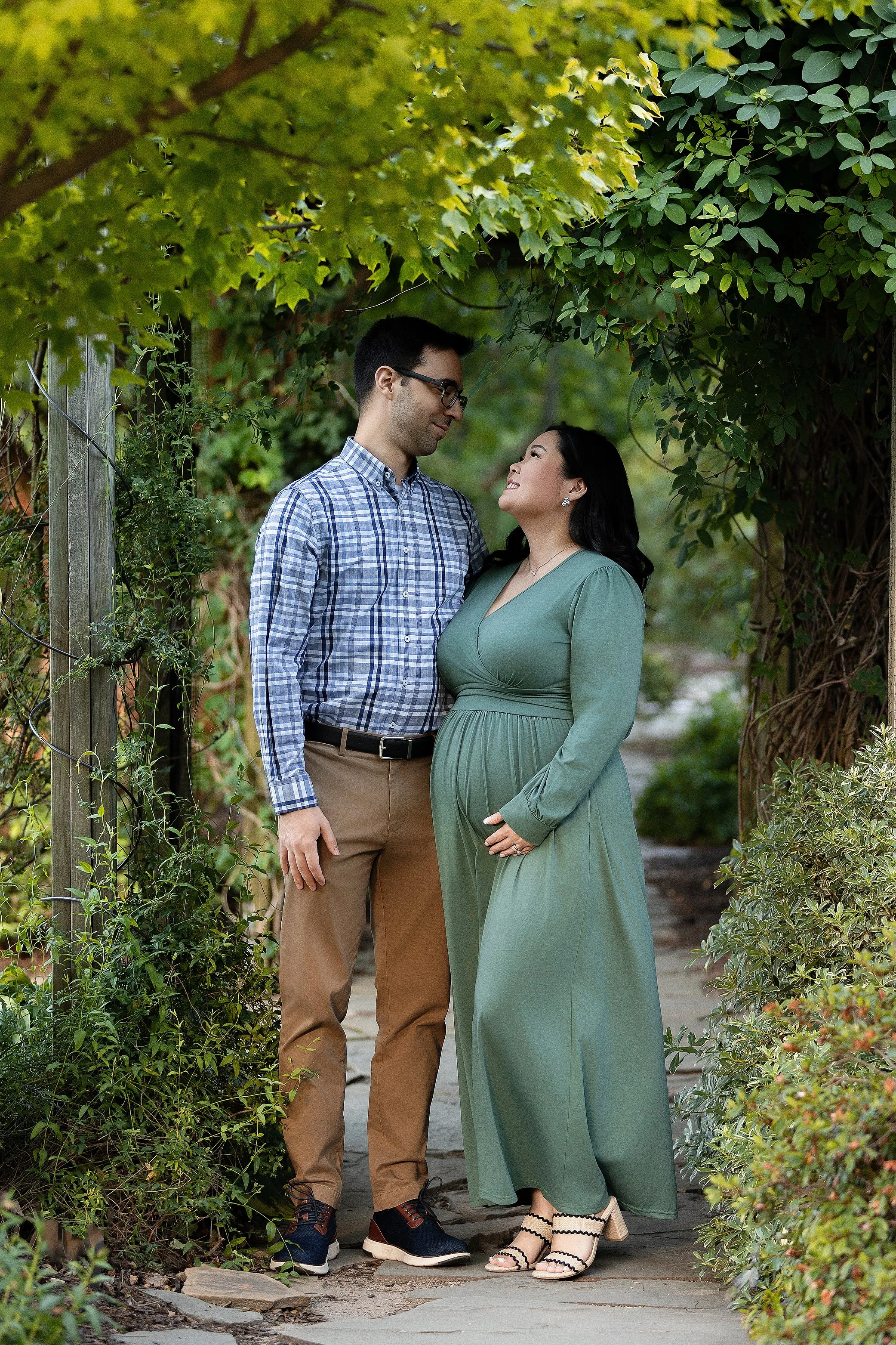 A man and woman standing close together on a garden path, smiling at each other beneath a leafy archway, with the woman holding her pregnant belly.