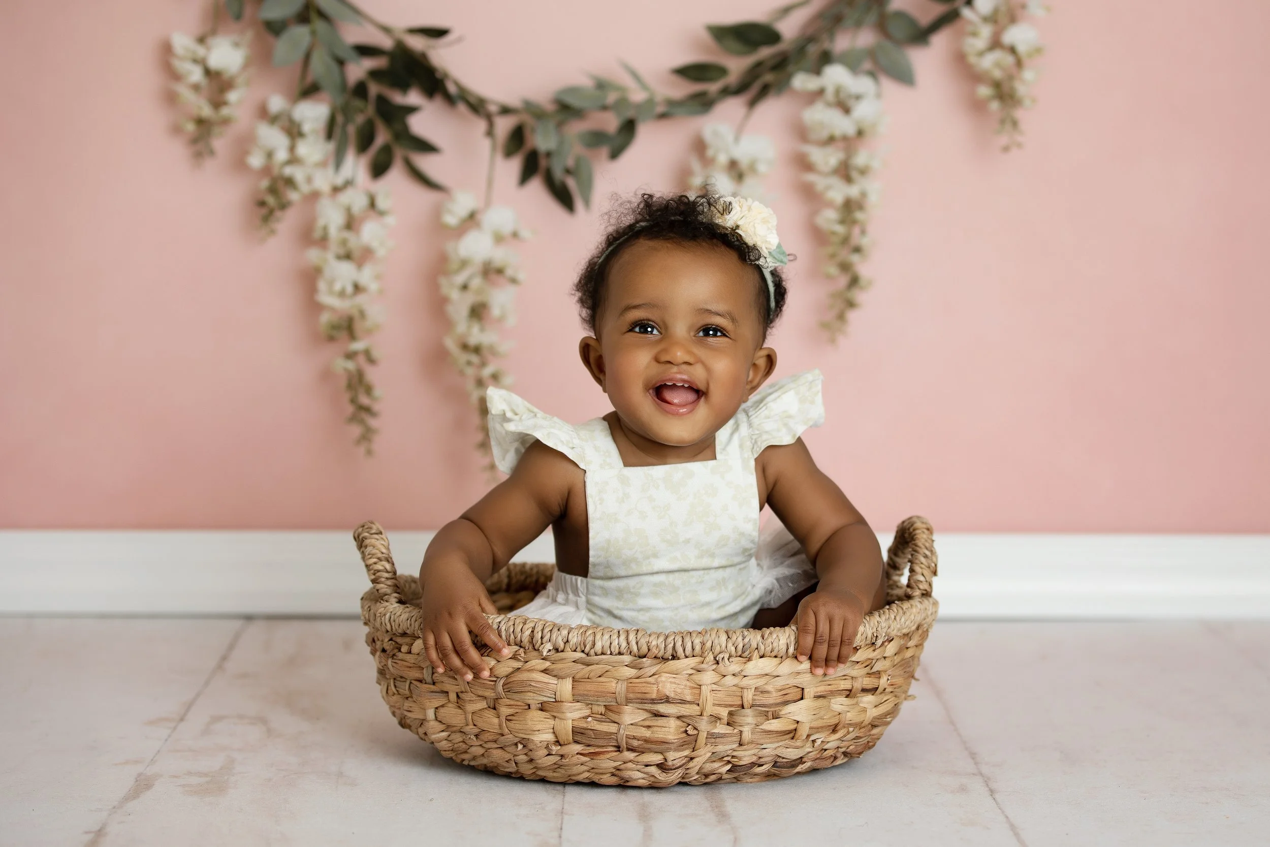 A smiling baby girl with curly hair, wearing a white floral dress and a flower headband, sitting in a woven basket against a pink wall decorated with hanging white flowers and green leaves.