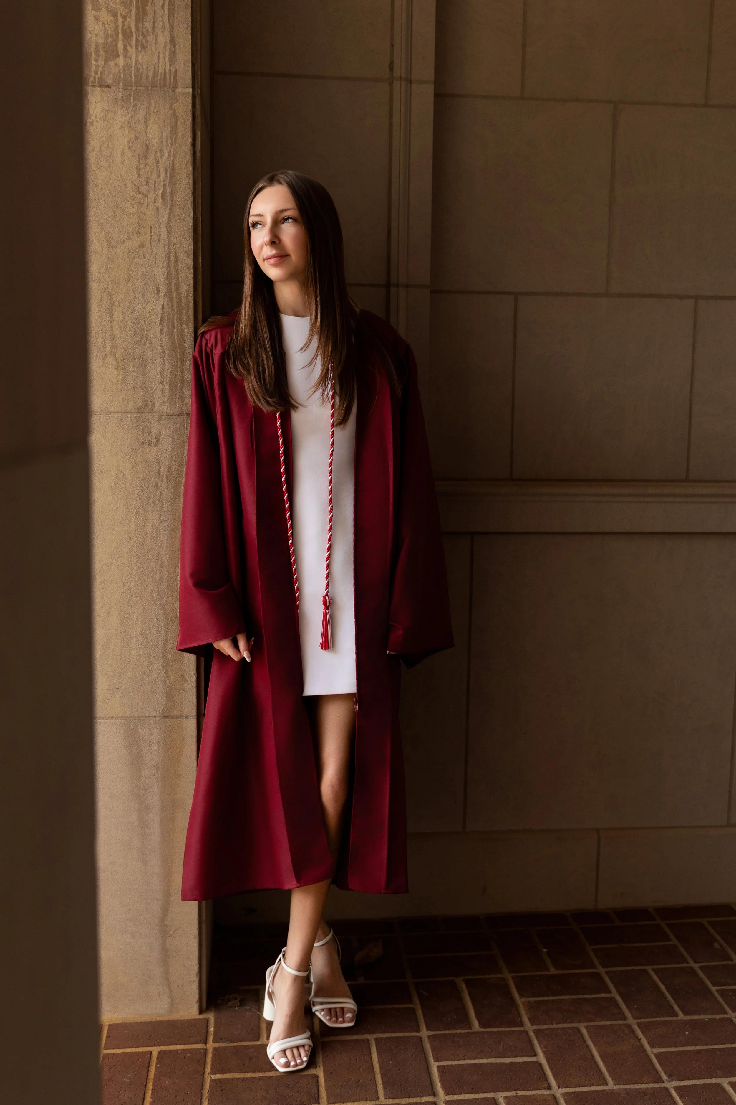 A young woman dressed in a white dress and wearing a maroon graduation gown with a red and white cord, standing near a wall with a contemplative expression, wearing white high heels.