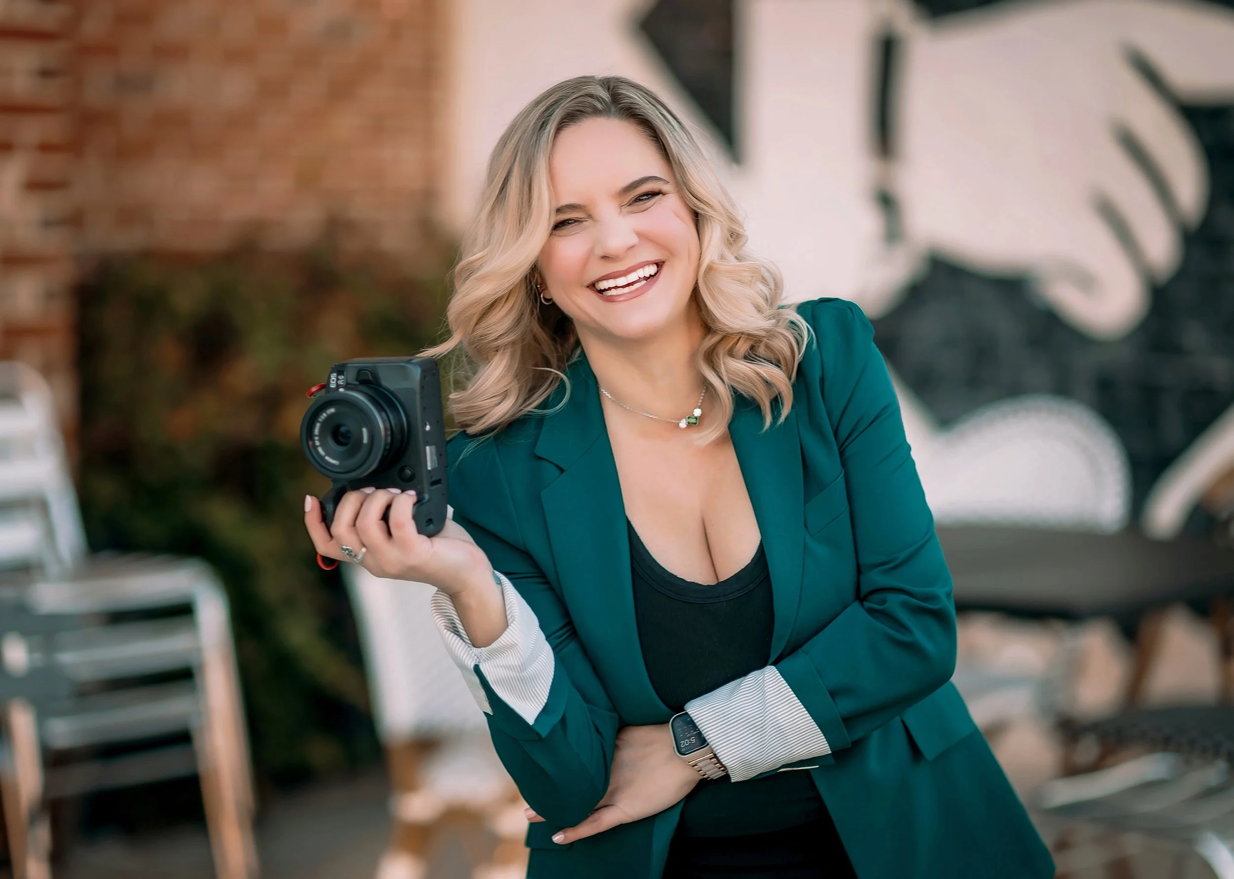 A smiling woman with blonde hair holding a camera outdoors.
