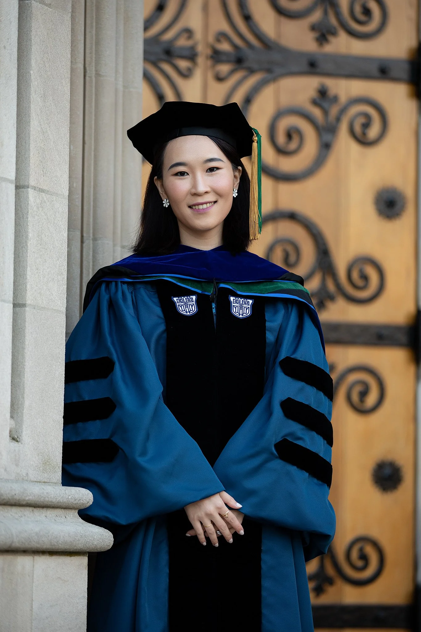 A young woman in a graduation cap and gown standing in front of a decorative wooden door with wrought iron details.