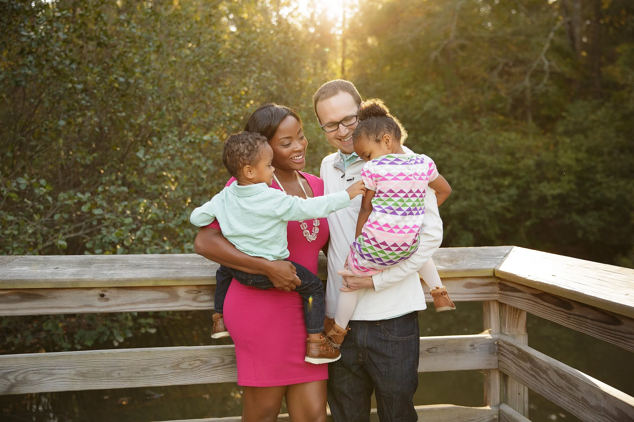 An African American woman, a Caucasian man, and two young children, one girl and one boy, enjoying a moment together outdoors during sunset on a wooden bridge surrounded by greenery.