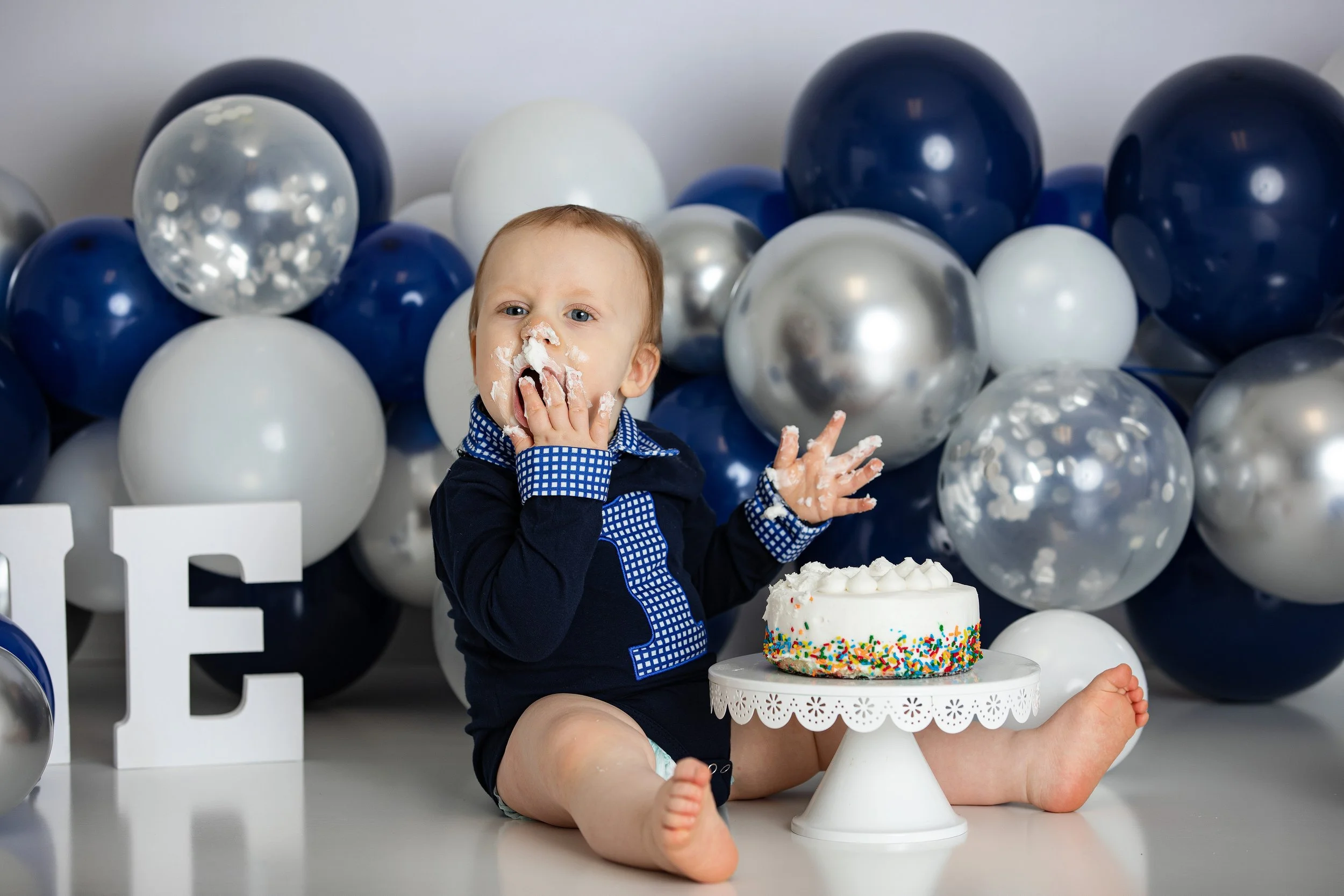 A young boy sitting on the floor celebrating a birthday with balloons in the background, a birthday cake with colorful sprinkles in front of him, and some balloons. The boy has cake on his nose and face, indicating he has been eating it.