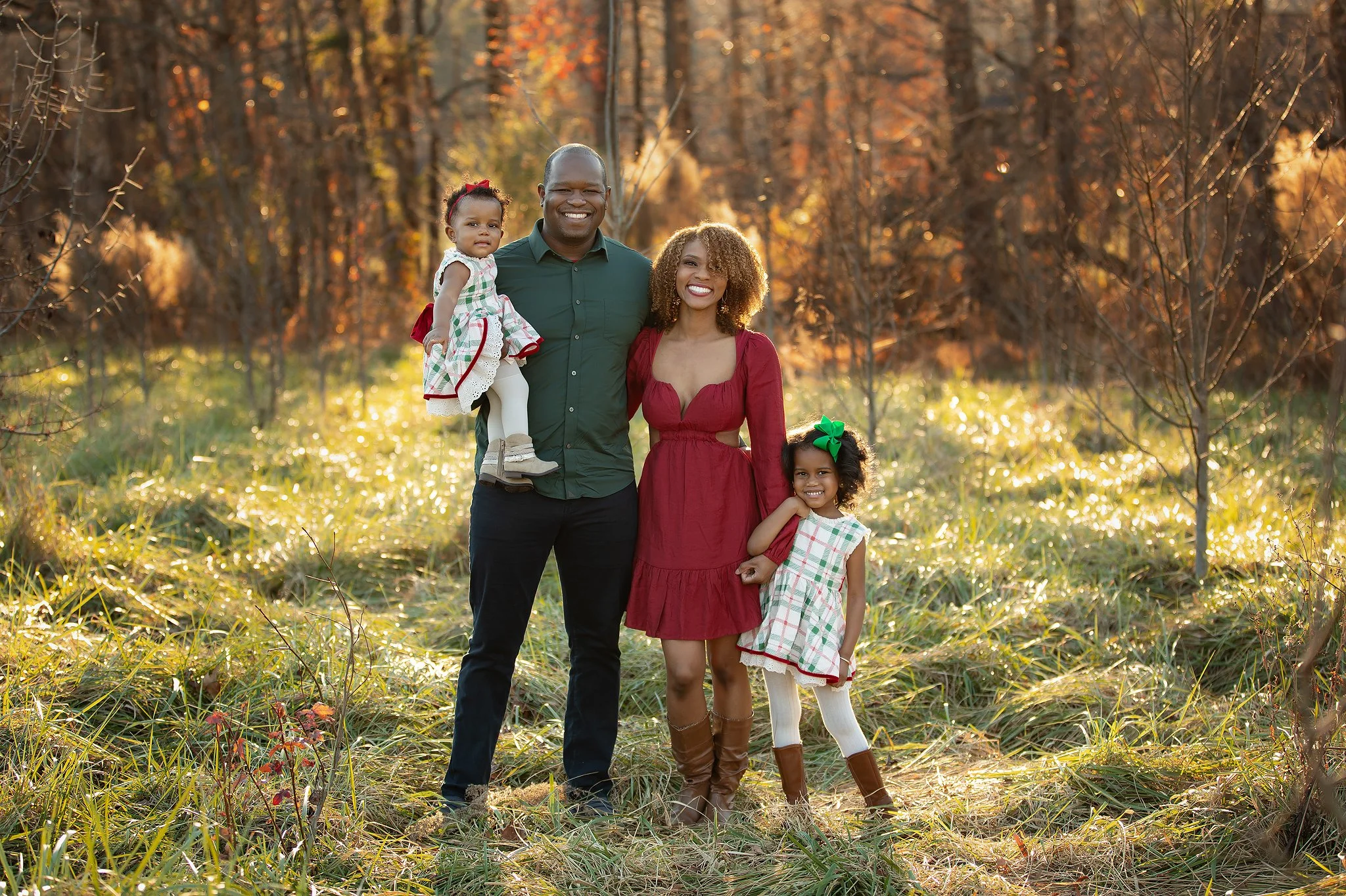 Family of four enjoying a fall day outdoors in a wooded area with colorful autumn leaves. The father is holding a young girl, the mother is smiling, and another young girl stands in front wearing a plaid dress and brown boots.