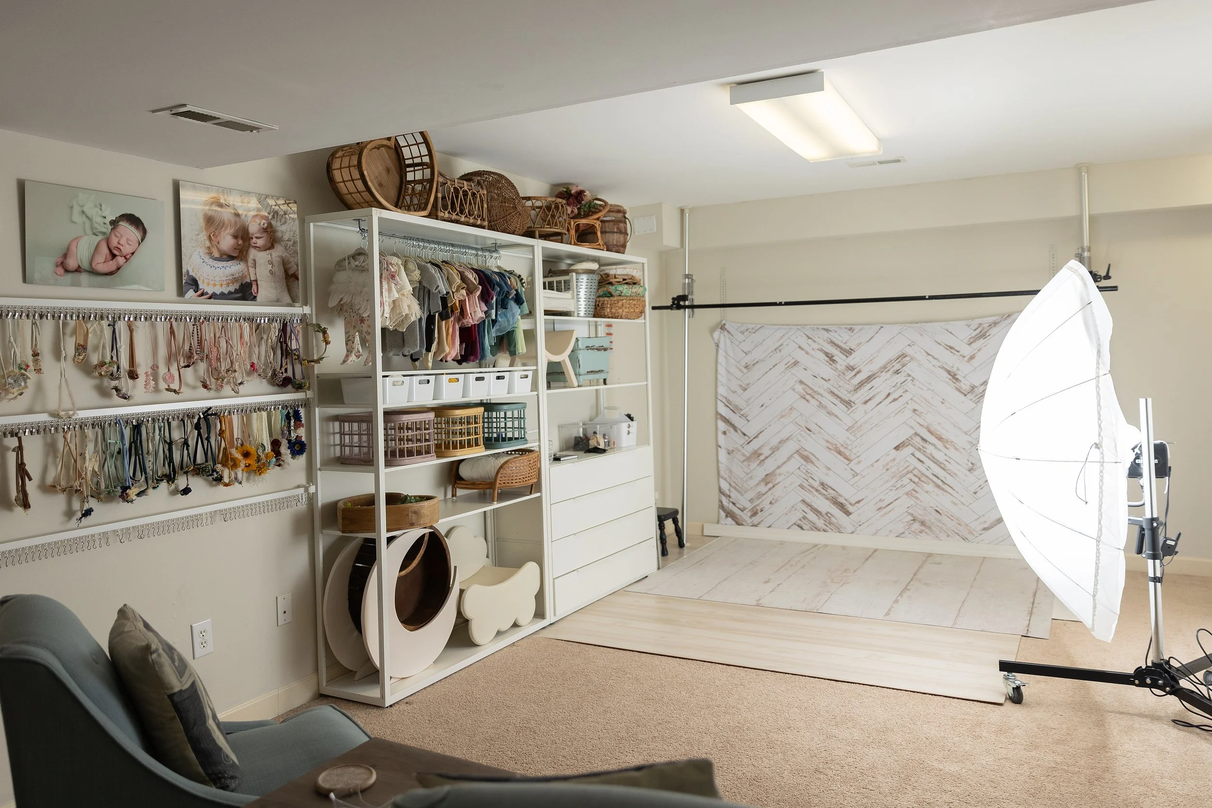 Photo studio setup with a white backdrop, professional lighting, and photography equipment, featuring shelves with children's clothing and decorative items, and walls adorned with children's artwork.