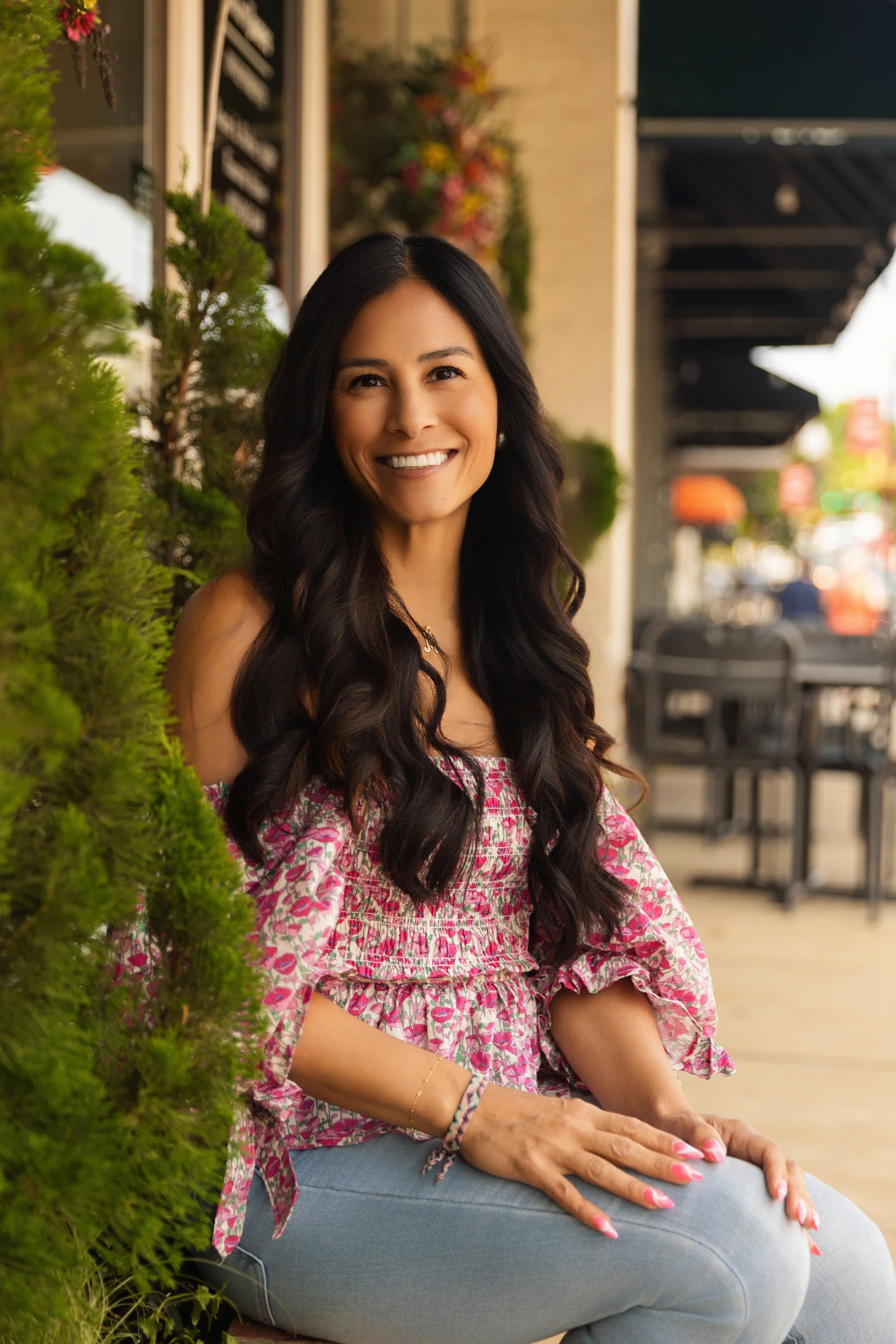 Smiling woman with long dark hair, sitting outdoors on a sidewalk, wearing a pink and white floral blouse and blue jeans, with greenery and a restaurant wall behind her.