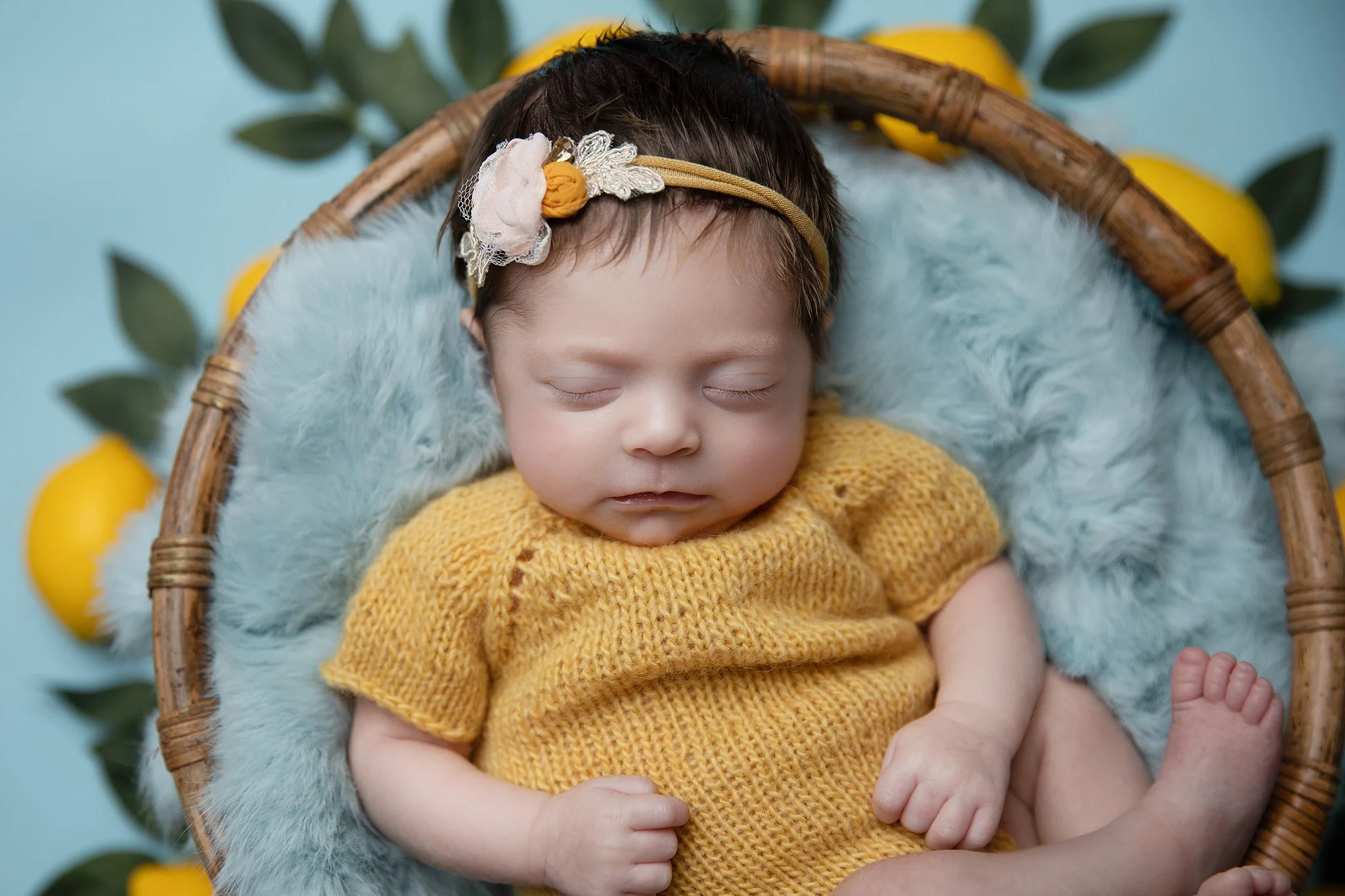 A sleeping baby with short dark hair, wearing a mustard yellow knitted outfit, lying in a round wicker basket with a fluffy white blanket and a decorative headband with fabric flowers. The background includes green leaves and yellow lemons.
