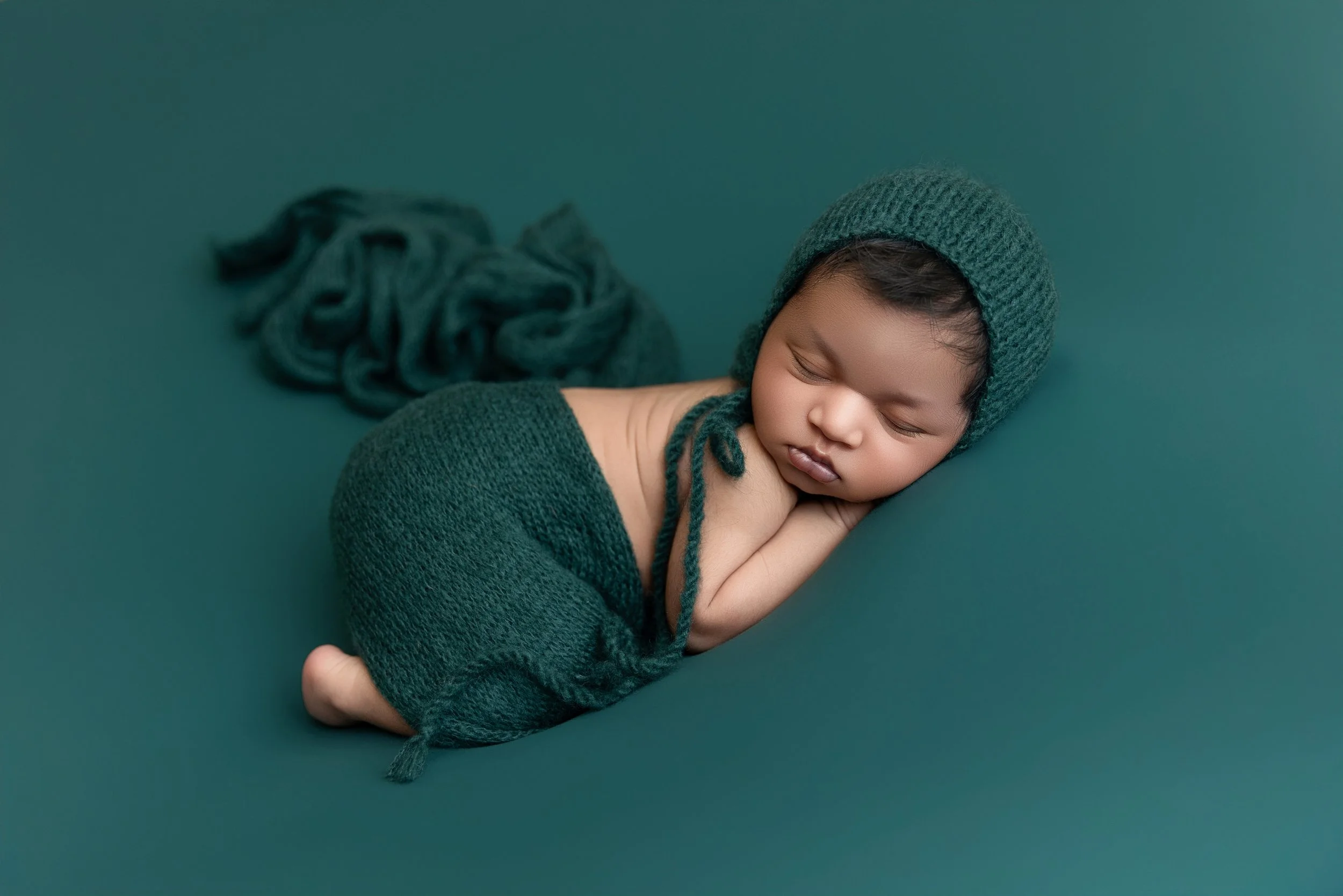 A sleeping child dressed in green, wearing a knitted hat and sweater, lying on a green surface with a pile of yarn in the background.