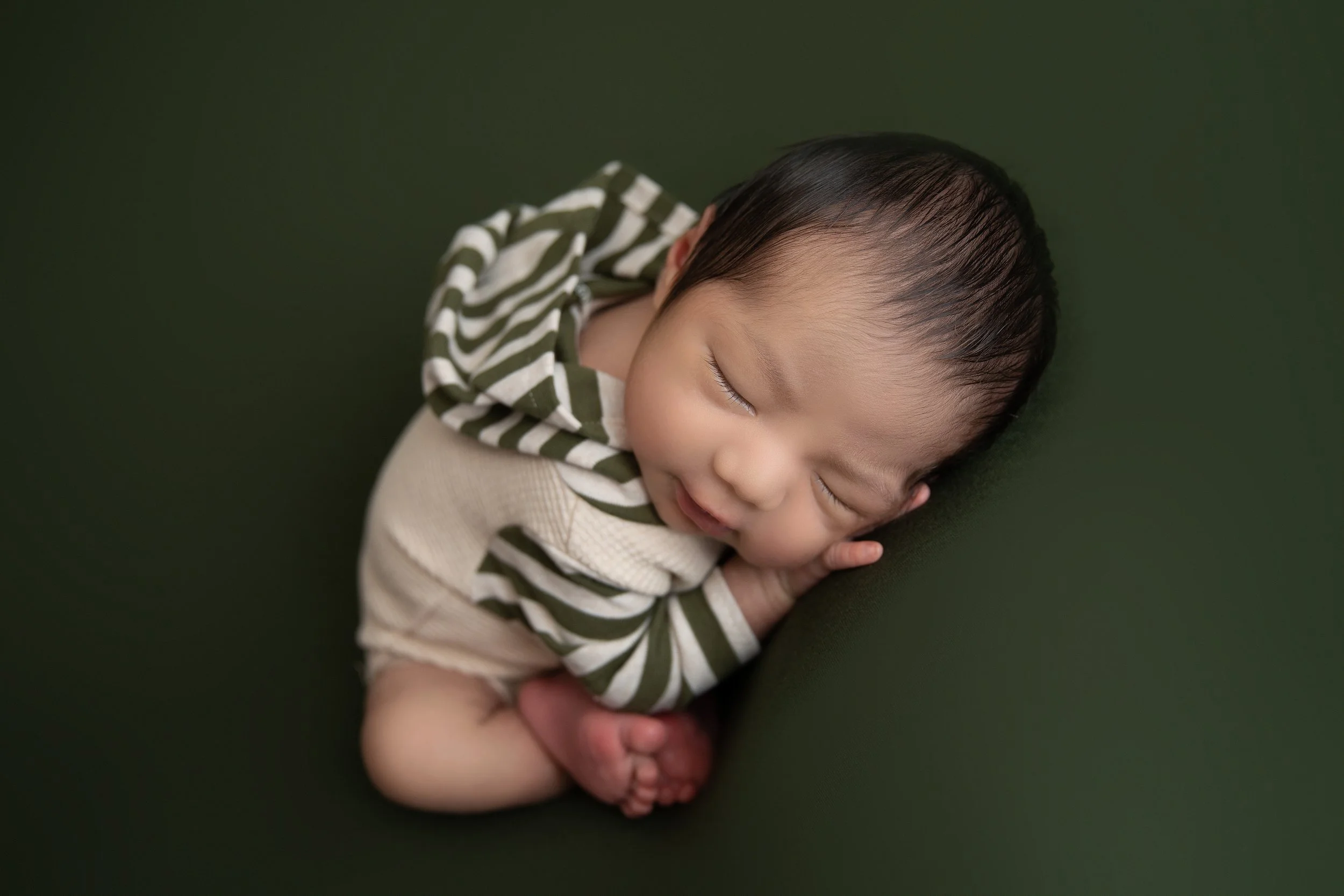 A young child with closed eyes sleeping, resting on a green surface, wearing a beige and green striped hoodie.