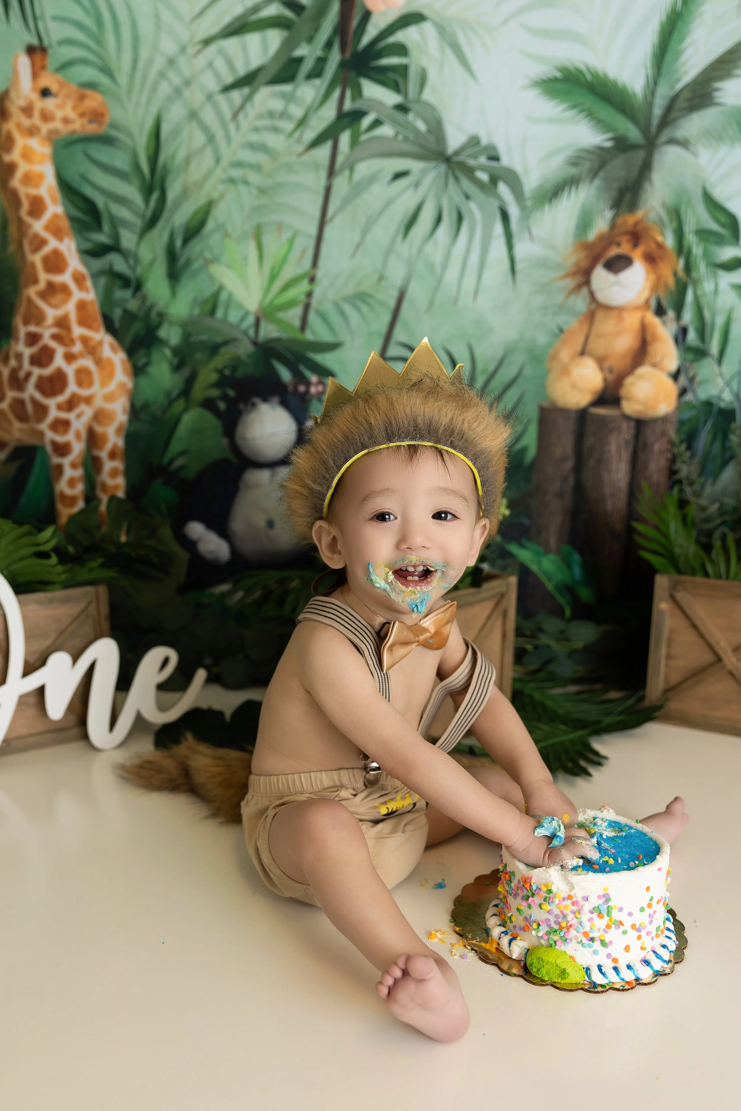 A young child with a lion mane costume and crown celebrating a birthday, sitting in front of a jungle-themed backdrop with plush animal toys and a decorated cake with blue frosting and colorful sprinkles.