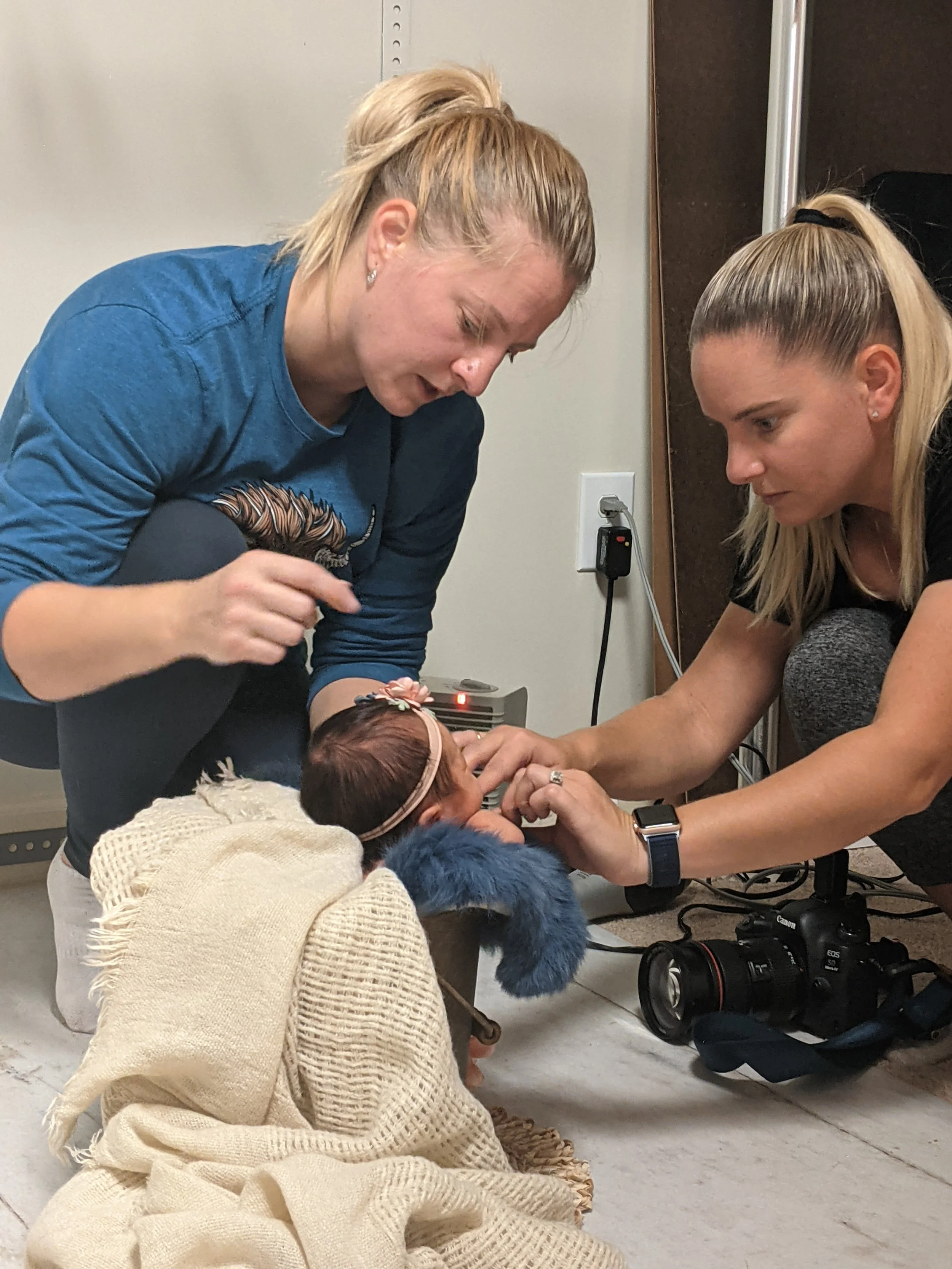 Two women are aiding a newborn baby in a hospital or birthing center, with one woman holding the baby's hand and the other adjusting the baby's head, surrounded by medical and camera equipment.