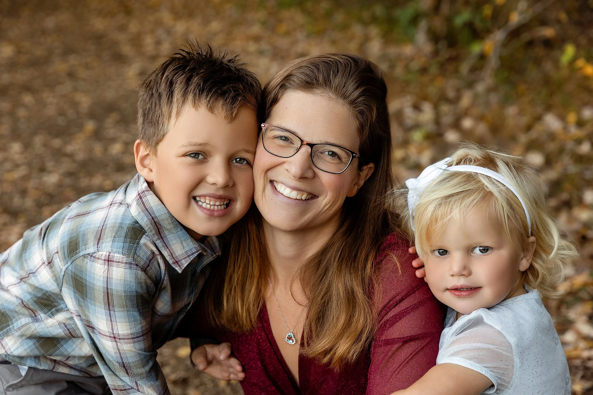A woman with glasses smiling and hugging two children, a boy and a girl, in an outdoor setting with autumn leaves on the ground.