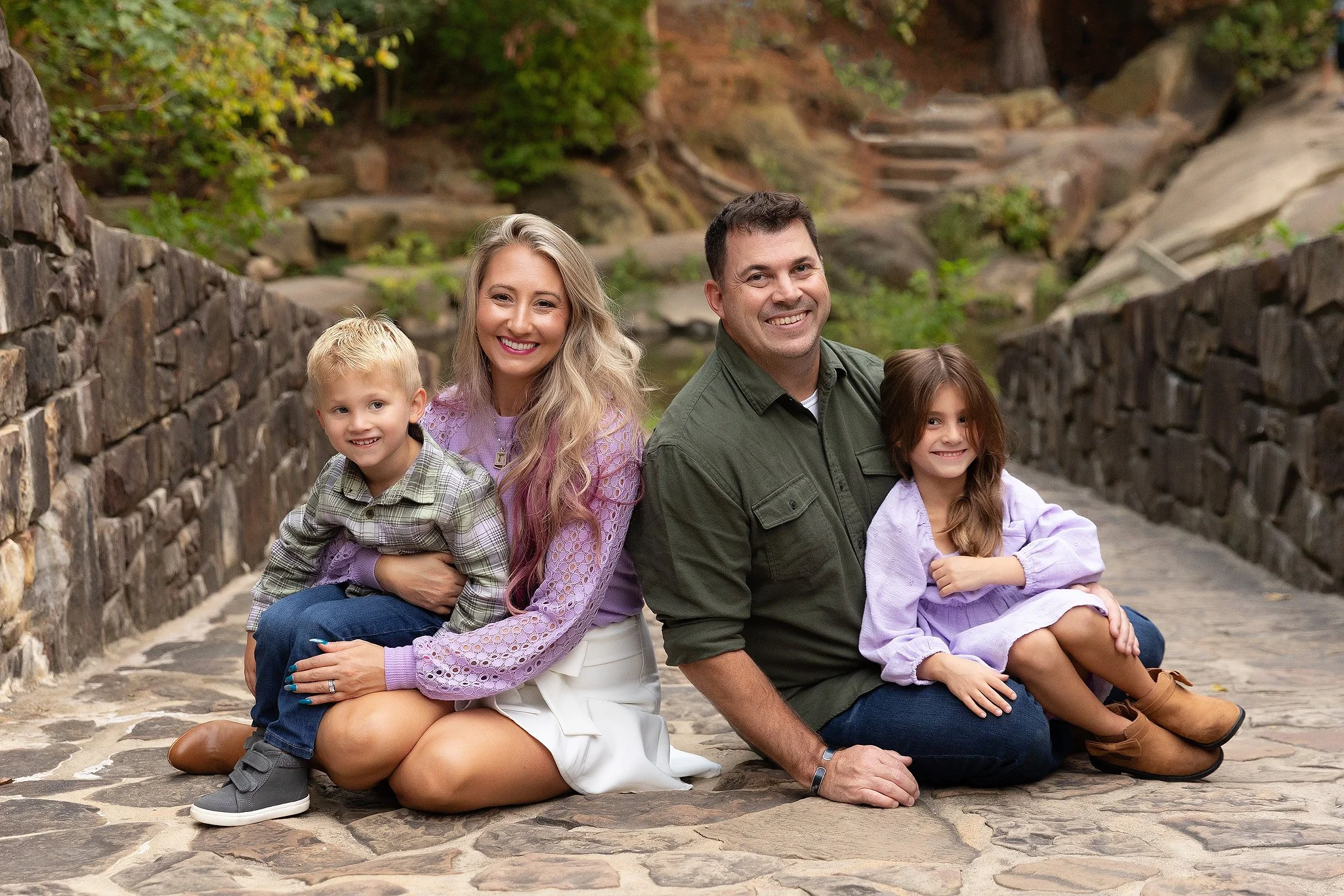 Family of five sitting together outdoors on a stone pathway beside a stone wall, smiling at the camera with trees and rocks in the background.