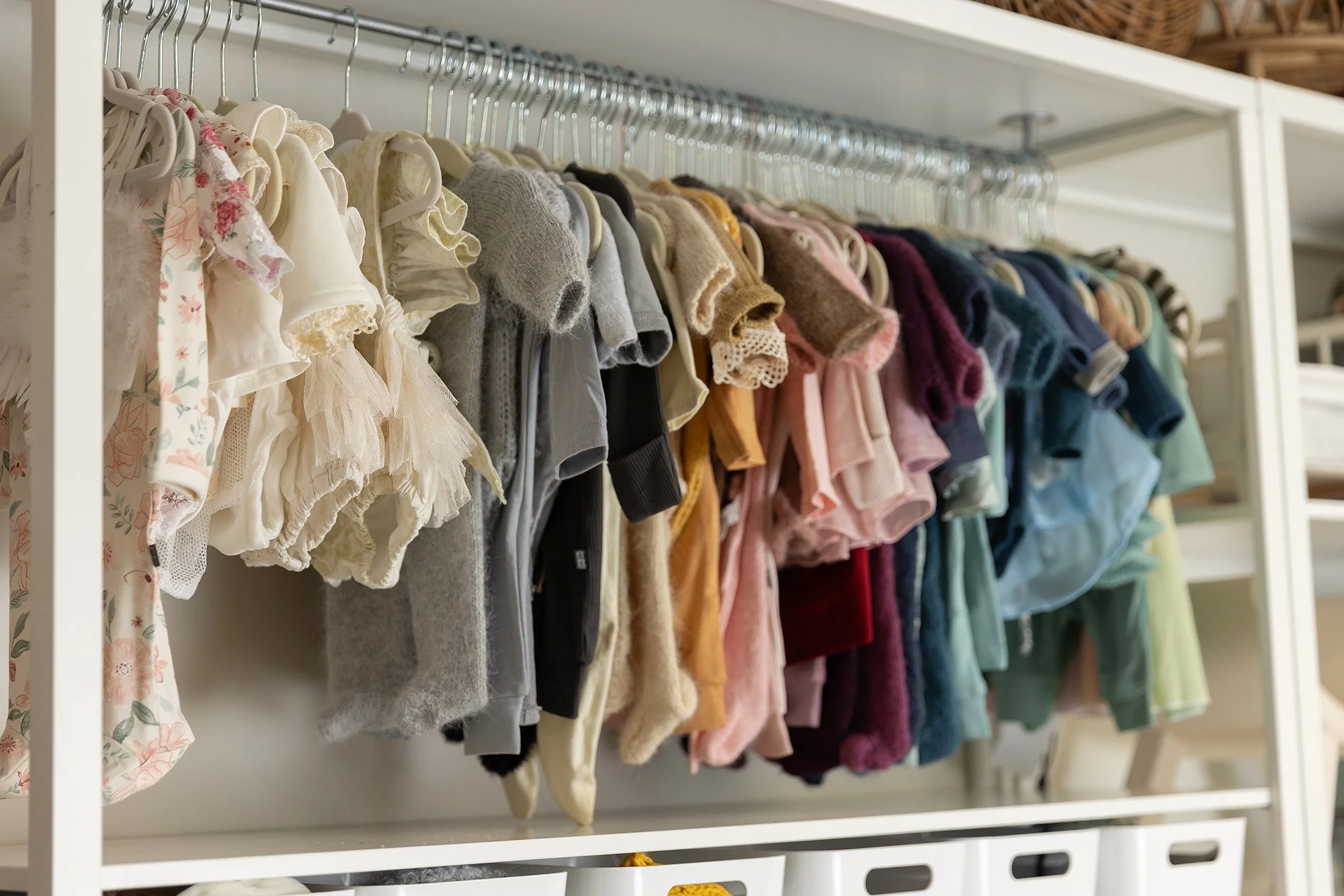 A white closet shelf with colorful baby clothes hanging on hangers, organized by color from light to dark.