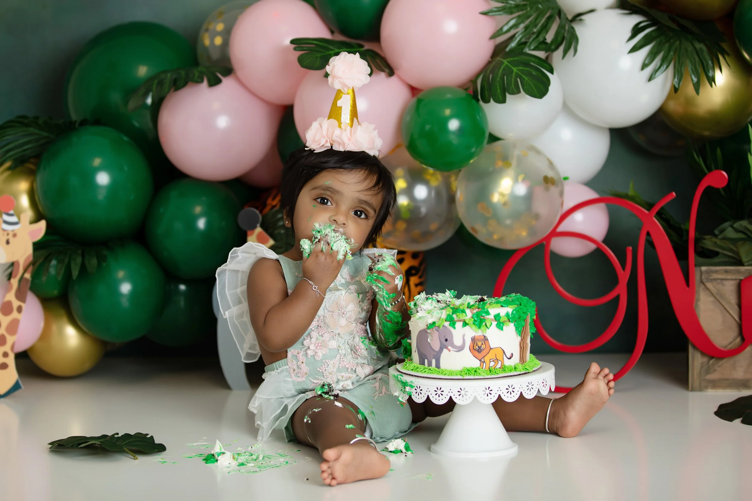 A young girl celebrating her first birthday, sitting in a high chair behind a decorated cake with animal illustrations, wearing a party hat with a gold '1' on it, surrounded by pink, green, and gold balloons and tropical leaves.