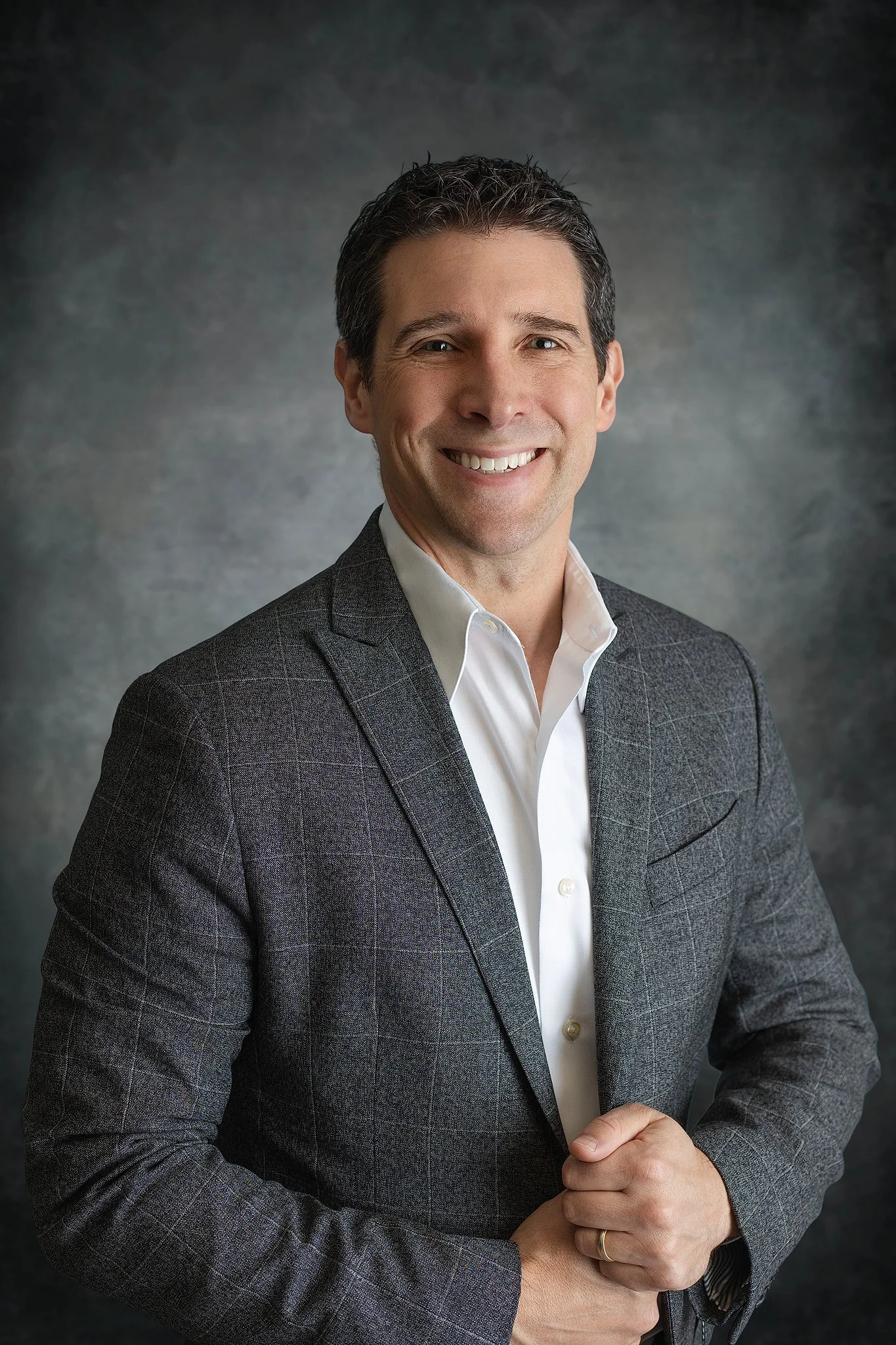 A man smiling, wearing a gray checkered suit and a white shirt, with his hands clasped in front of him, against a gray textured background.
