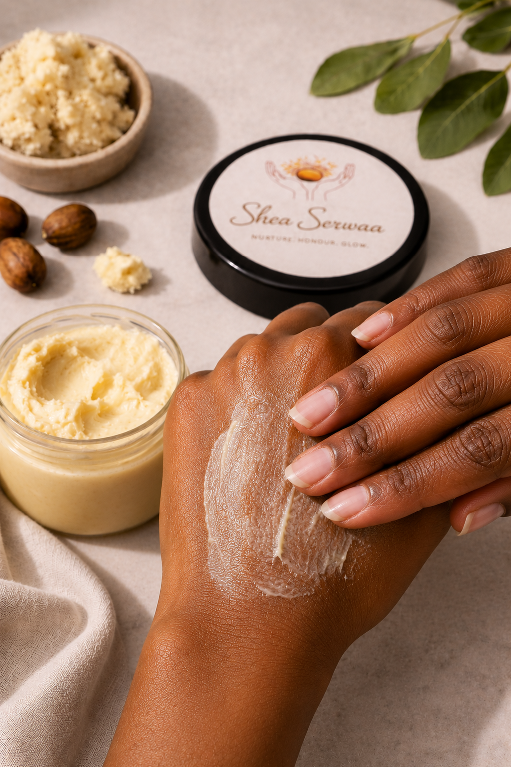A person applying Shea Sewaa moisturizer to their hand. The scene includes a jar of shea butter, a bowl of crushed shea nuts, a container of shea butter, and a black container labeled Shea Sewaa. Green leaves are in the background.