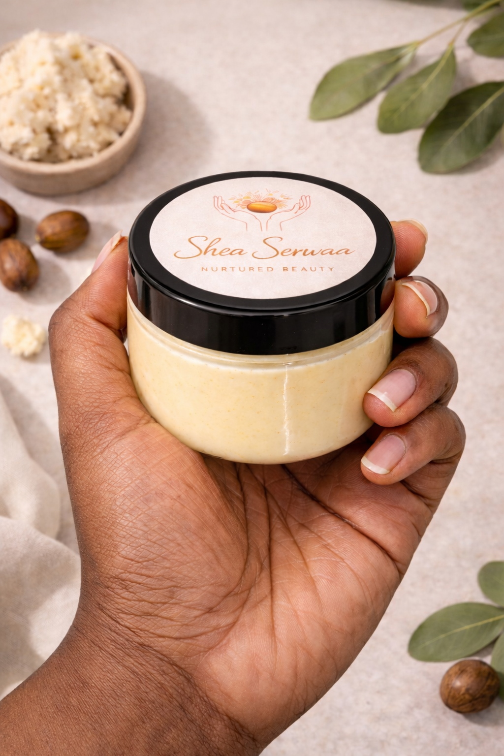 A hand holding a jar of Shea Serwaa skincare product with a black lid and white label, surrounded by shea nuts, a small bowl of shea butter, and green leaves on a light surface.