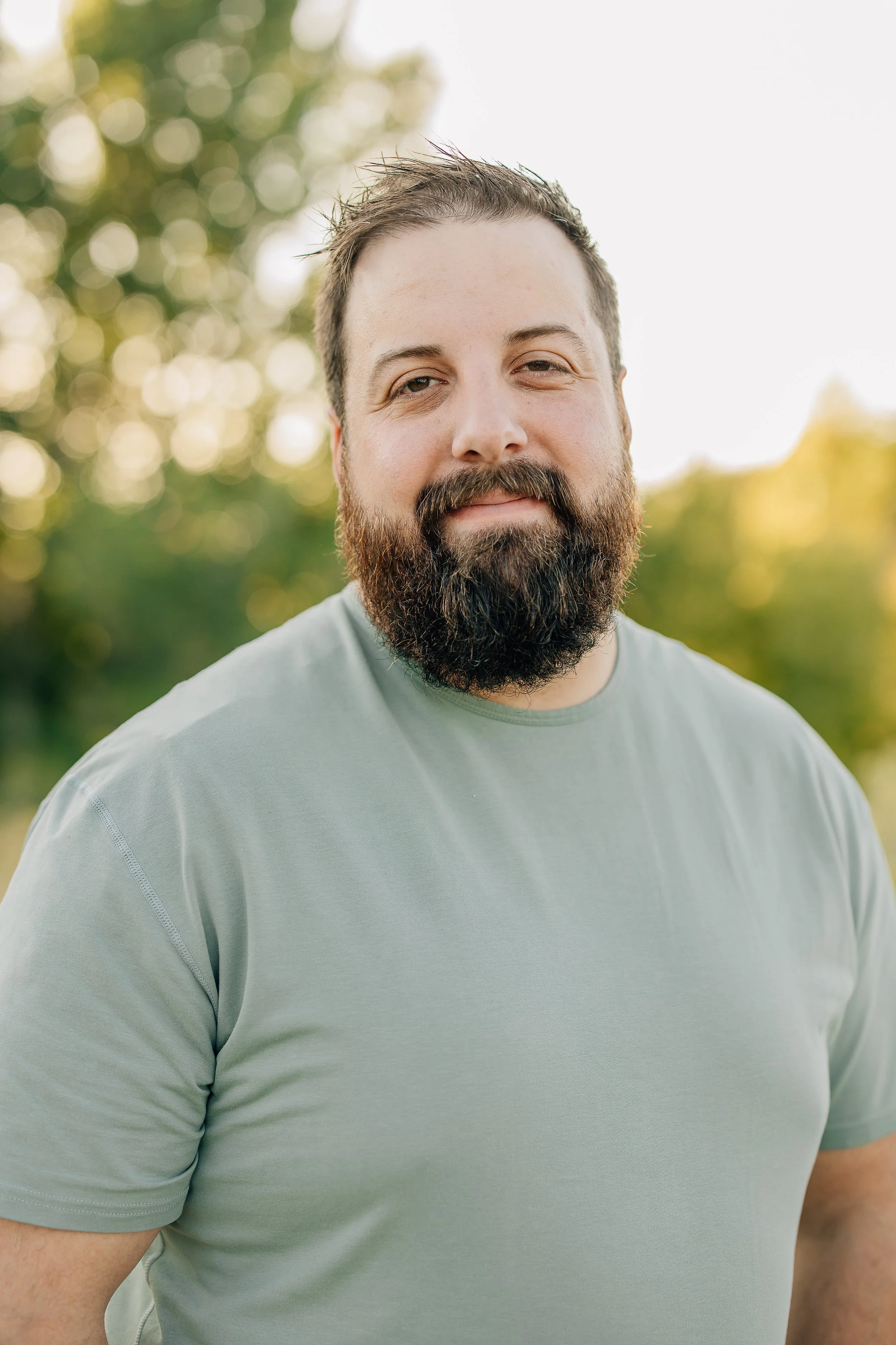 Close-up of a man with a beard and short hair wearing a light green t-shirt standing outdoors with blurred trees in the background.