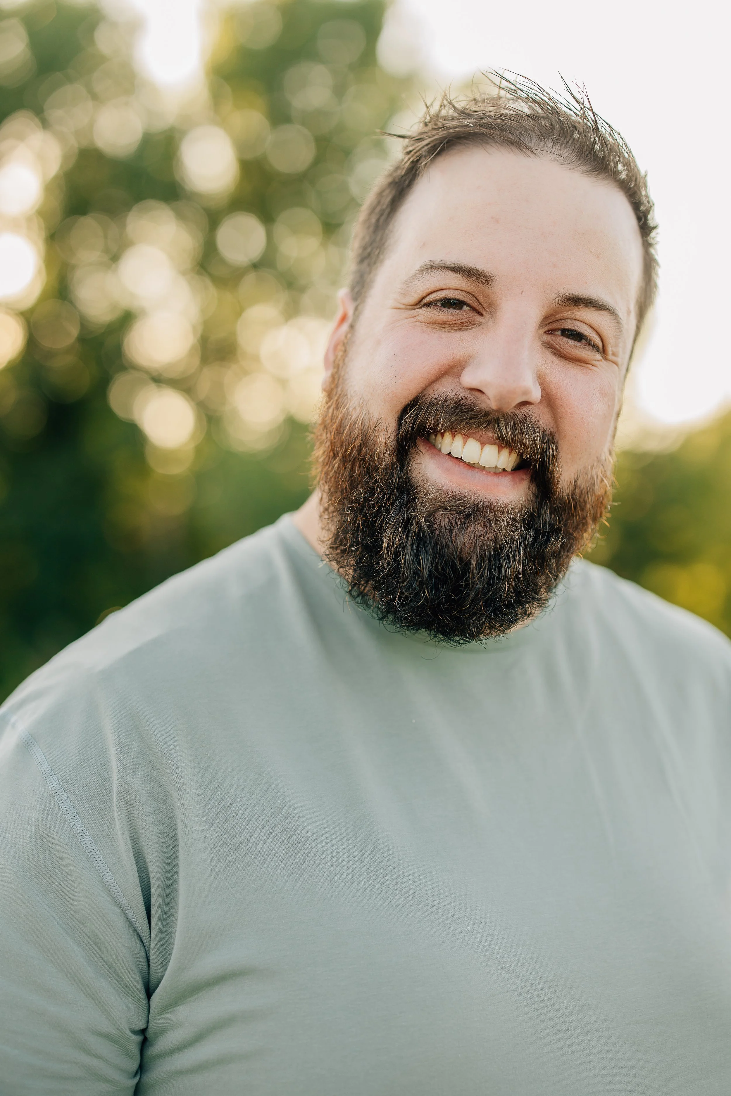 Close-up of a man with a beard smiling outdoors with sunlight and blurred green background.