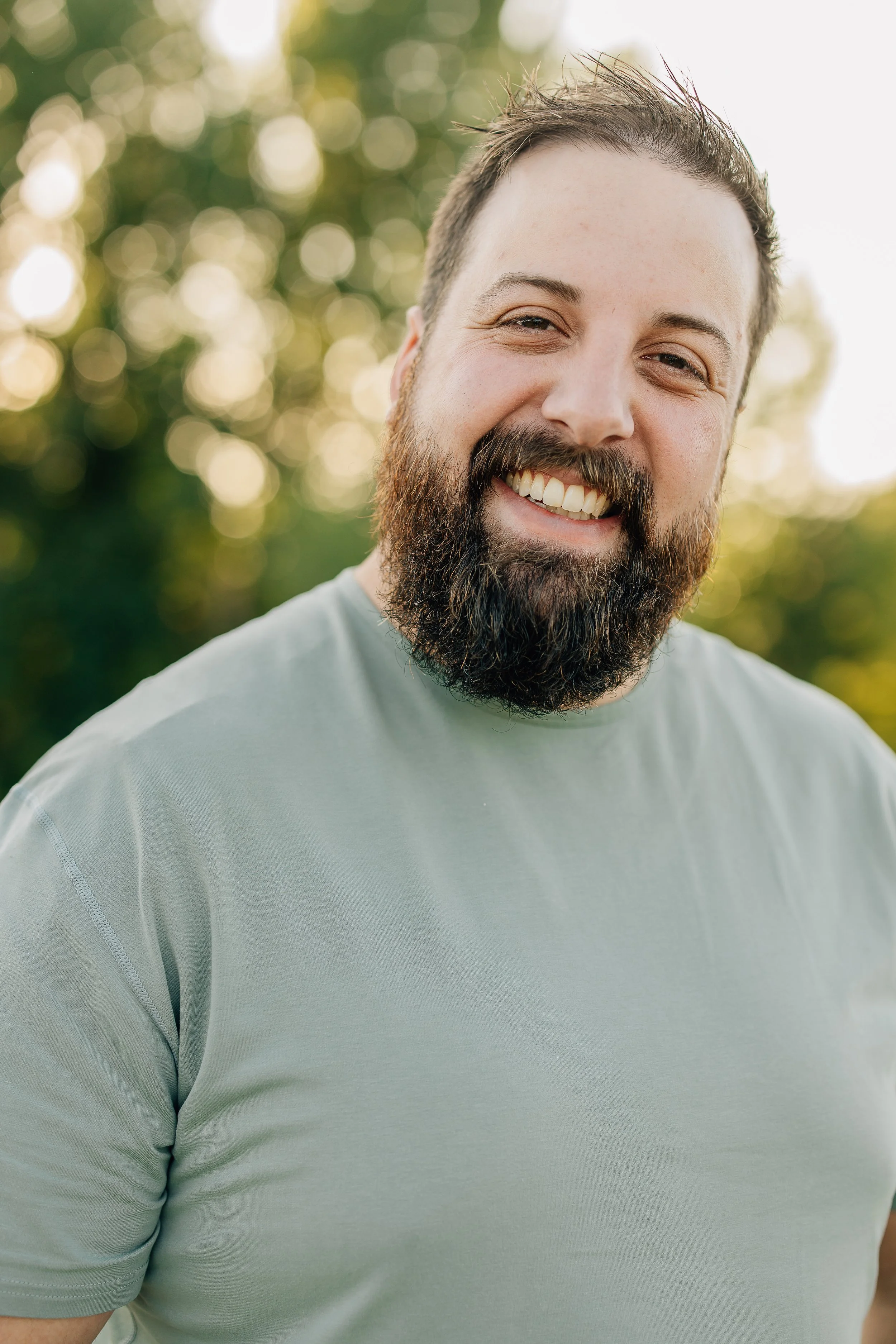 Close-up of a smiling man with a beard and short dark hair, wearing a light gray T-shirt outdoors with blurred green foliage in the background.