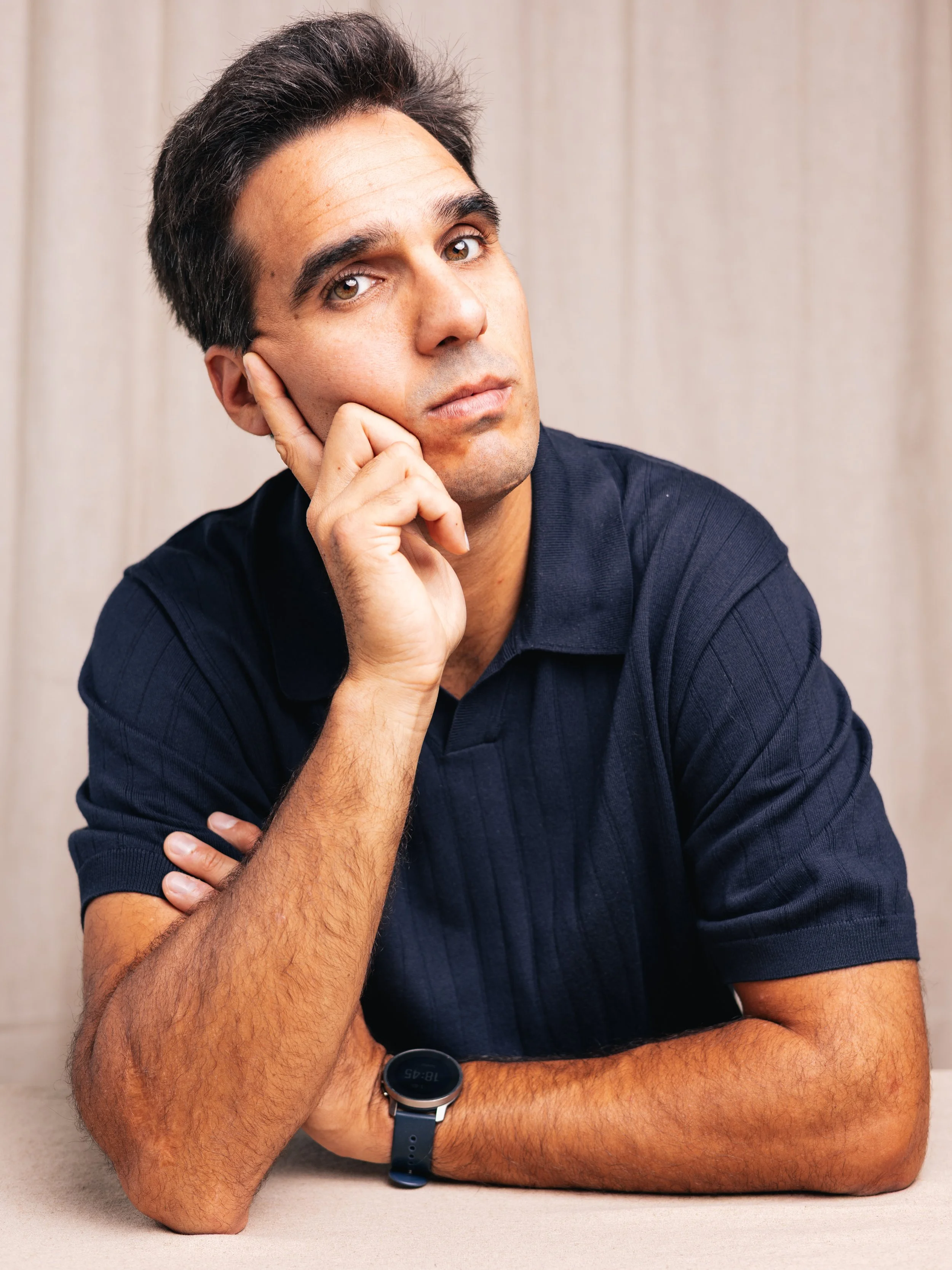 A man with short dark hair and brown eyes, wearing a black shirt and a smartwatch, sitting at a table with his left arm resting on the surface. He is resting his head on his left hand and looking directly at the camera with a neutral expression. The background is a plain beige wall.