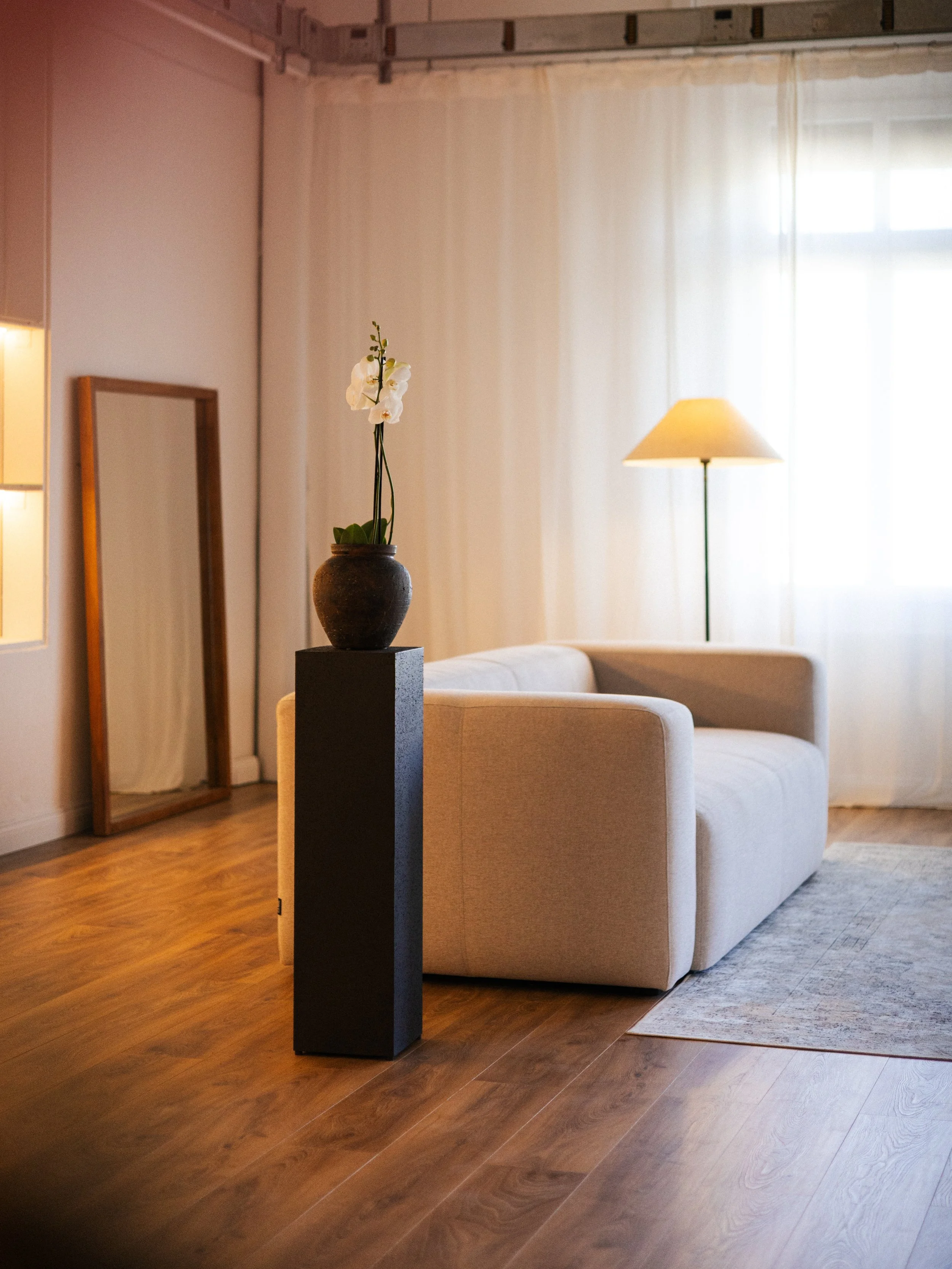 Living room with beige sofa, wooden flooring, a black pedestal with a potted white orchid, a floor lamp, a mirror, and a large window with white curtains.
