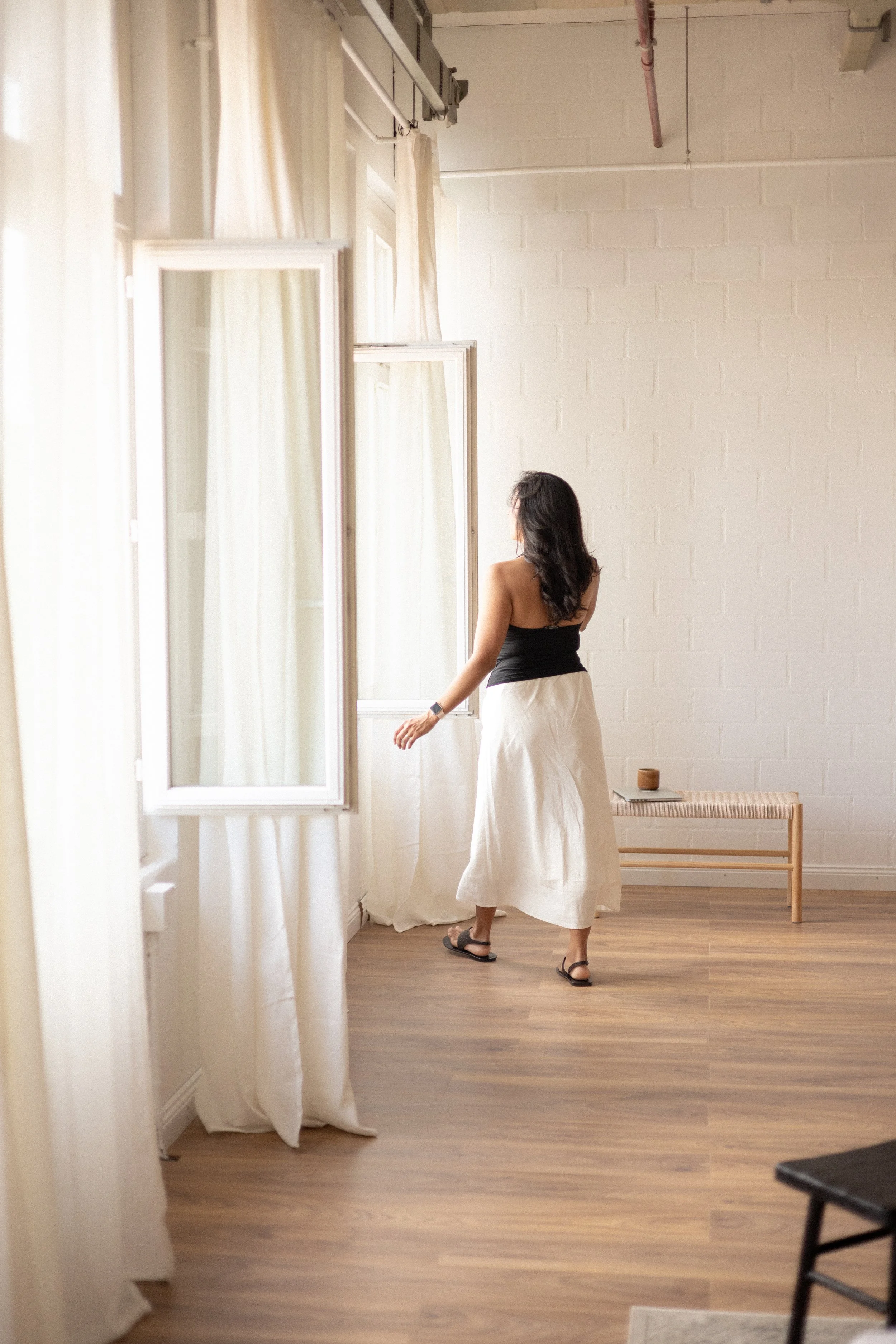A woman with dark hair wearing a black top, a white skirt, and sandals, standing in a bright room, looking out the window, with a small wooden table with a candle on it in the background.