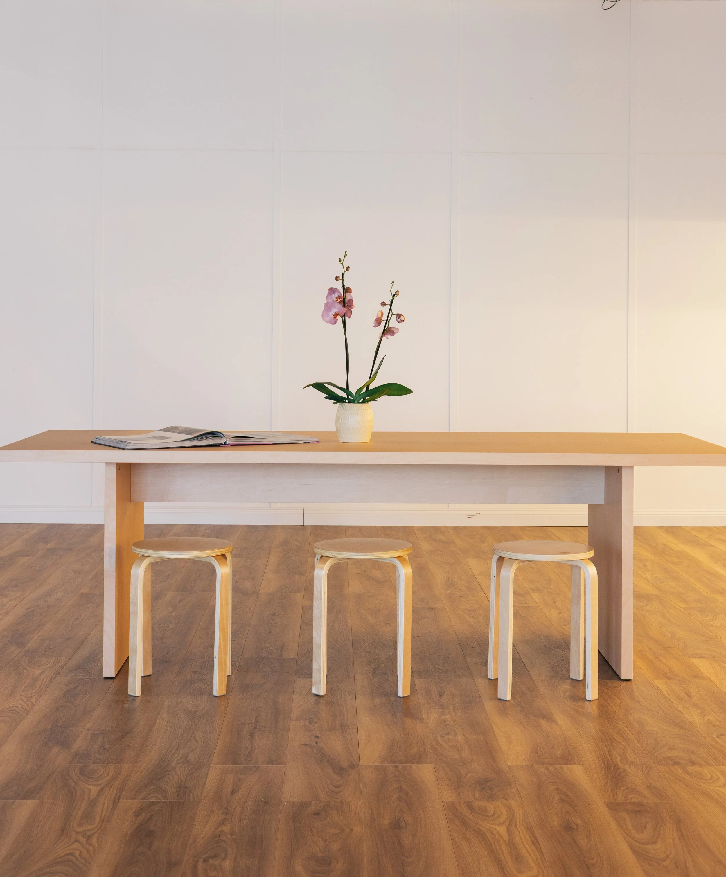 A wooden table with three stools and a pink orchid in a white vase in a minimalist room with white tiled walls.