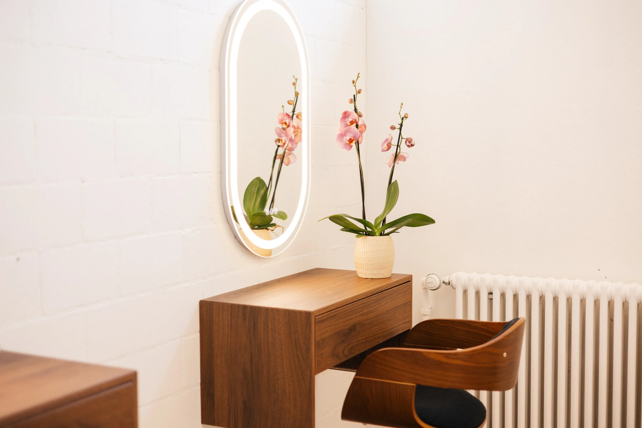 A wooden desk with a small white orchid plant in a beige woven pot, a mirror with a white frame reflecting the orchid, and a white radiator on a white brick wall.