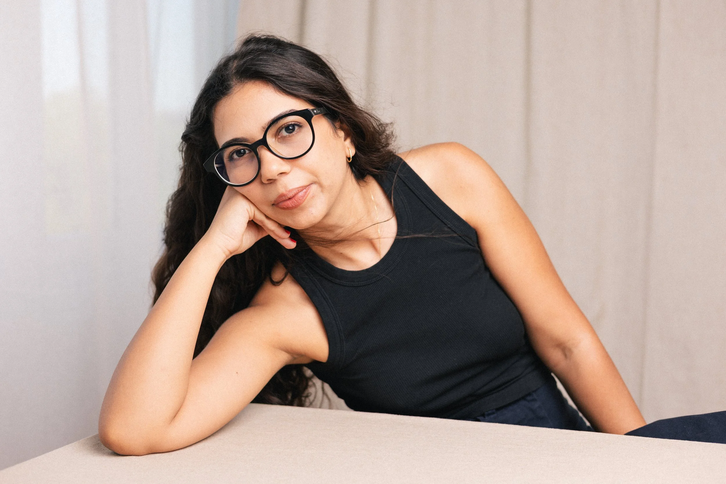 A woman with long dark hair, wearing black glasses and a black sleeveless top, rests her chin on her hand while sitting at a beige table with a neutral background.