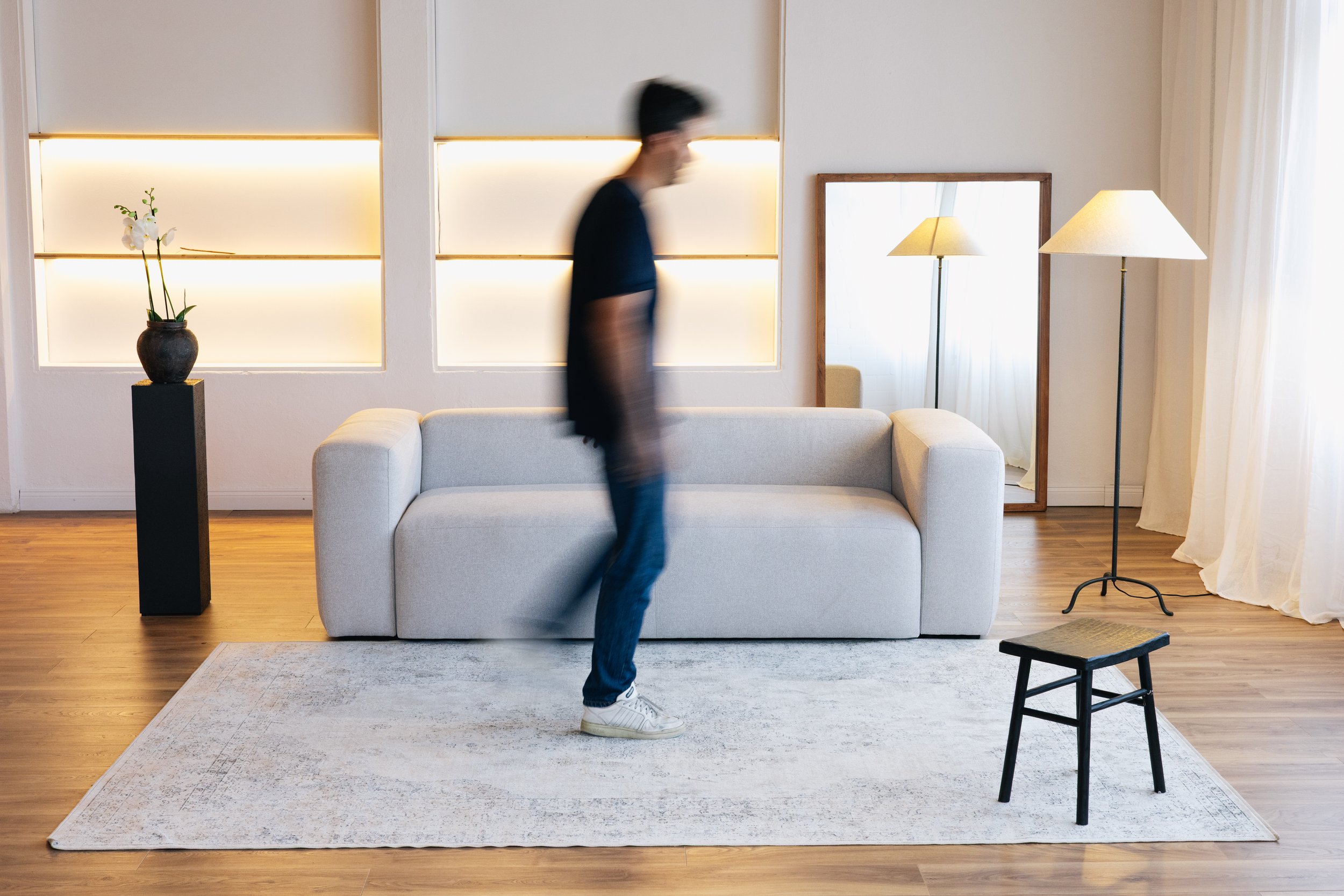 A person walking in a living room with a beige sofa, a large mirror, and two standing lamps. The room has wooden flooring, a white rug, a large window with white curtains, and a black stool in the foreground.
