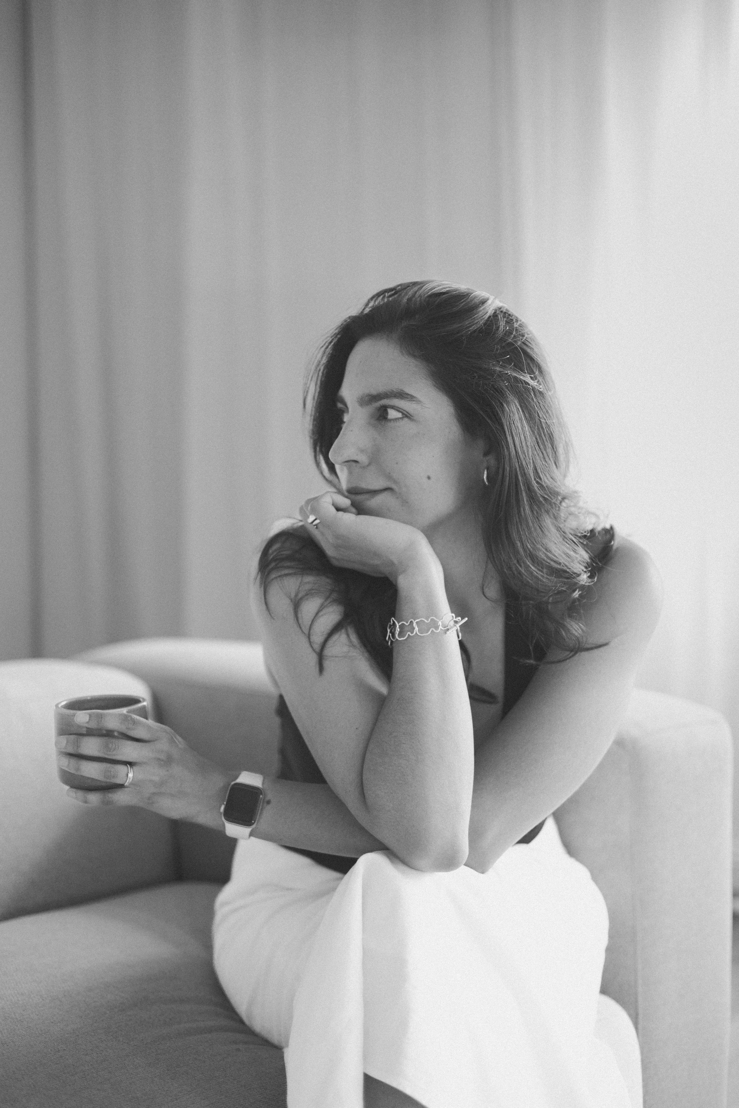 A woman with dark hair and earrings is sitting on a sofa, holding a cup of coffee with her right hand, while her left arm rests on her knee. She is looking to her right with her chin resting on her hand, wearing a watch and a bracelet. The background features a light-colored curtain.