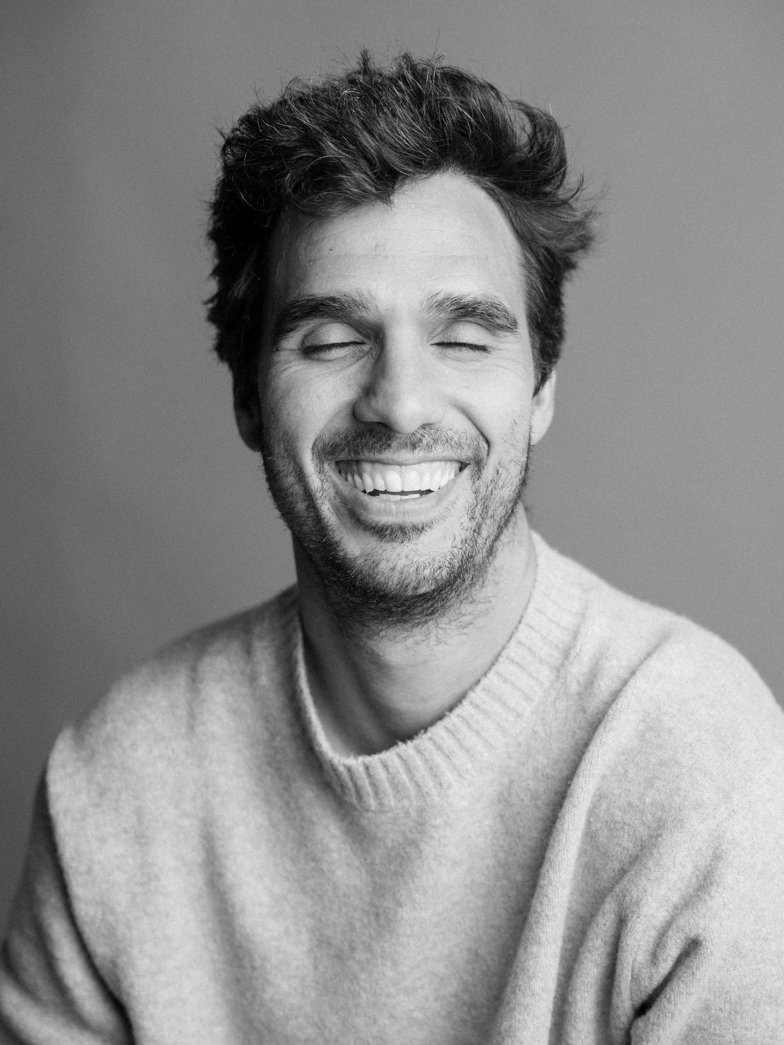 Black and white portrait of a man smiling with closed eyes, wearing a light-colored sweater.