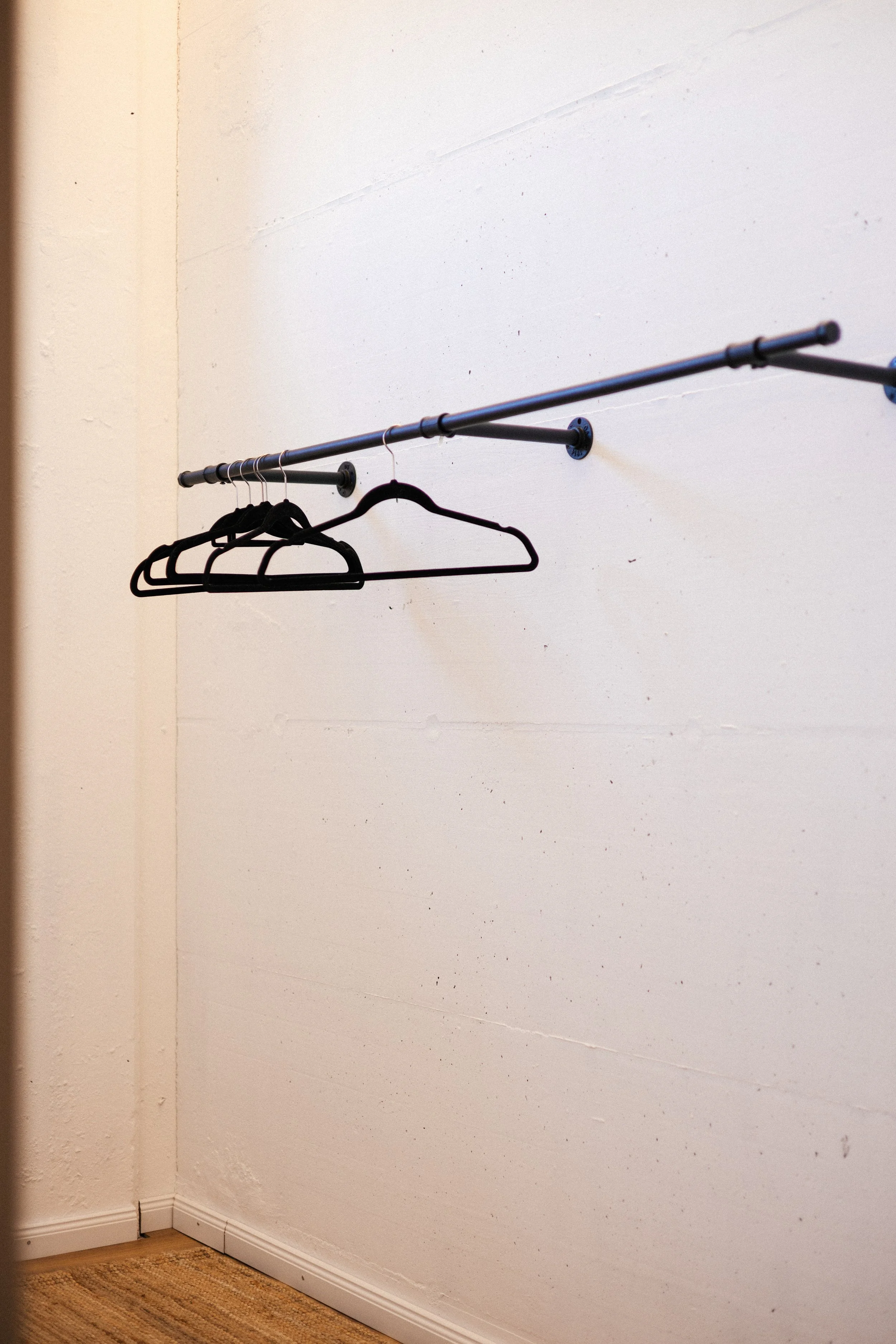 Empty black coat and clothing hangers on a black metal rod mounted on a white wall in a room with wooden flooring and a corner with baseboard.