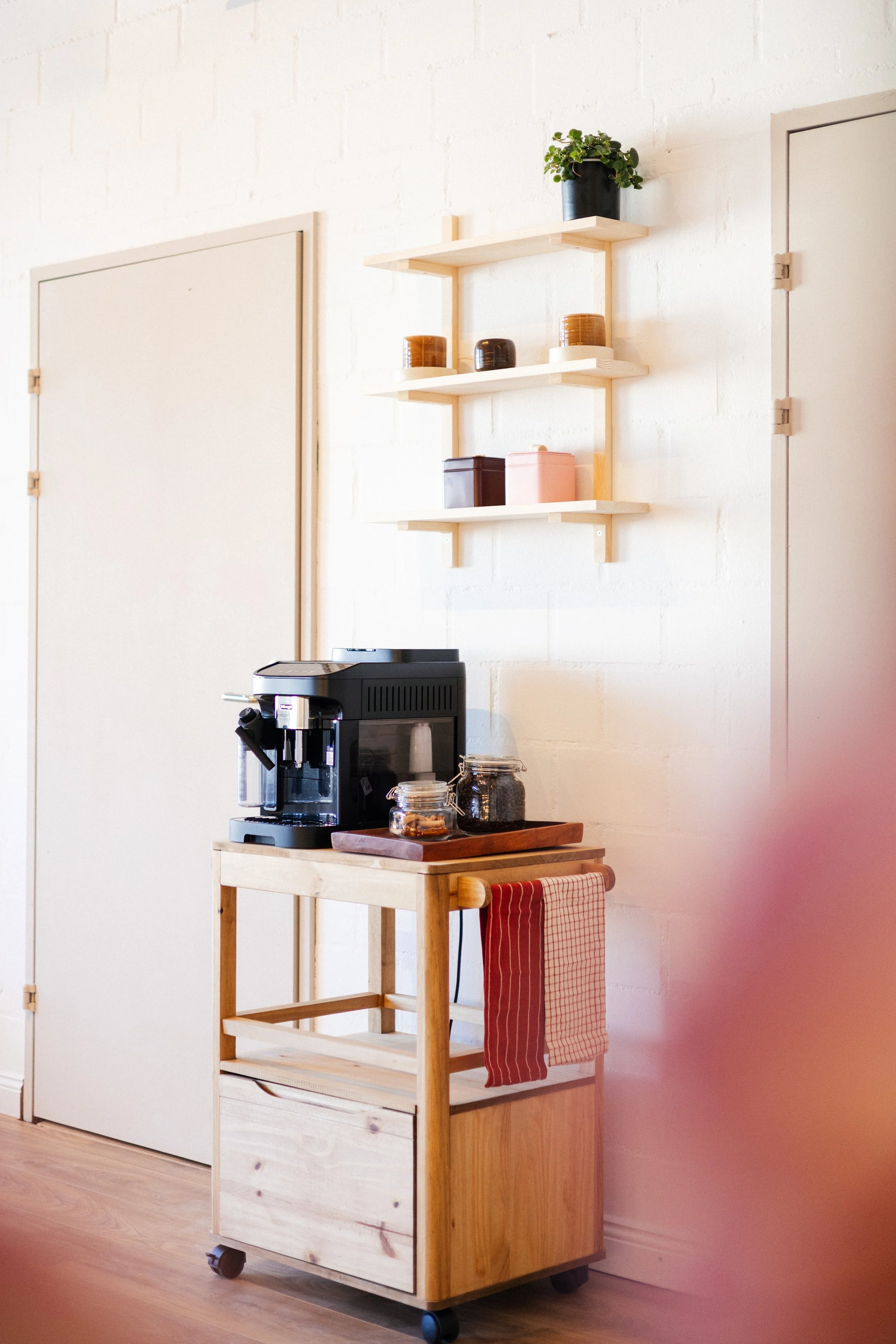 A small wooden rolling cart with a coffee machine on top, a tray, and jars of coffee beans, positioned against a white wall. Behind it, a wooden shelf with decorative jars and a potted plant is mounted on the wall.