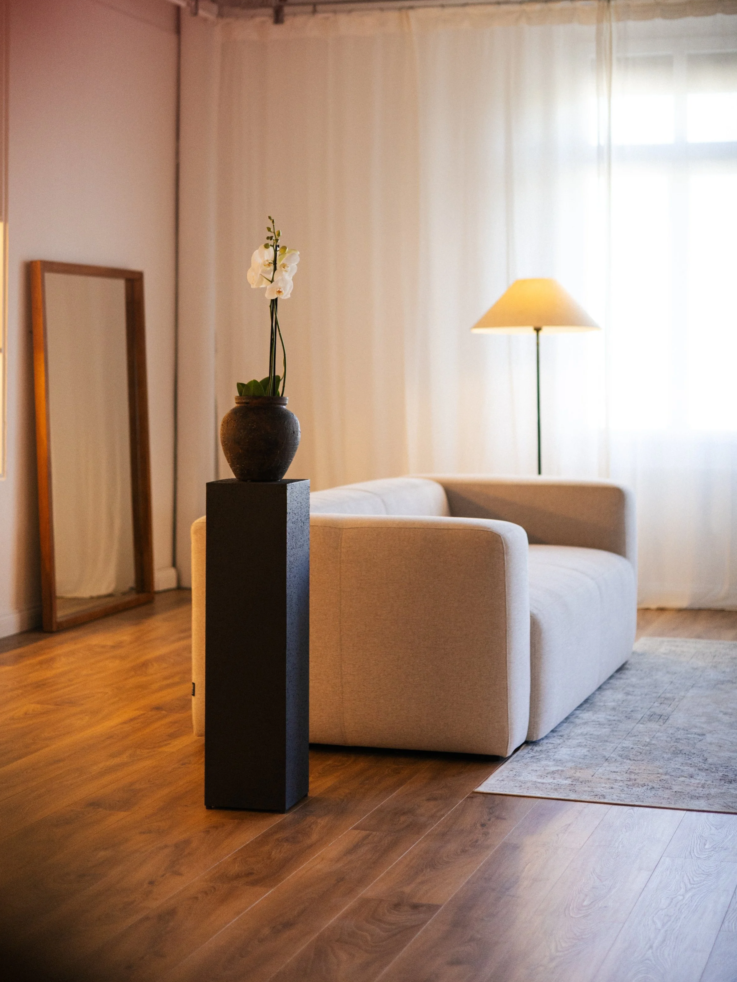Living room with a white sofa, a wooden framed mirror leaning against the wall, a floor lamp with a yellow shade, a black pedestal with a small orchid plant in a round brown pot, hardwood floor, and sheer white curtains covering a window.