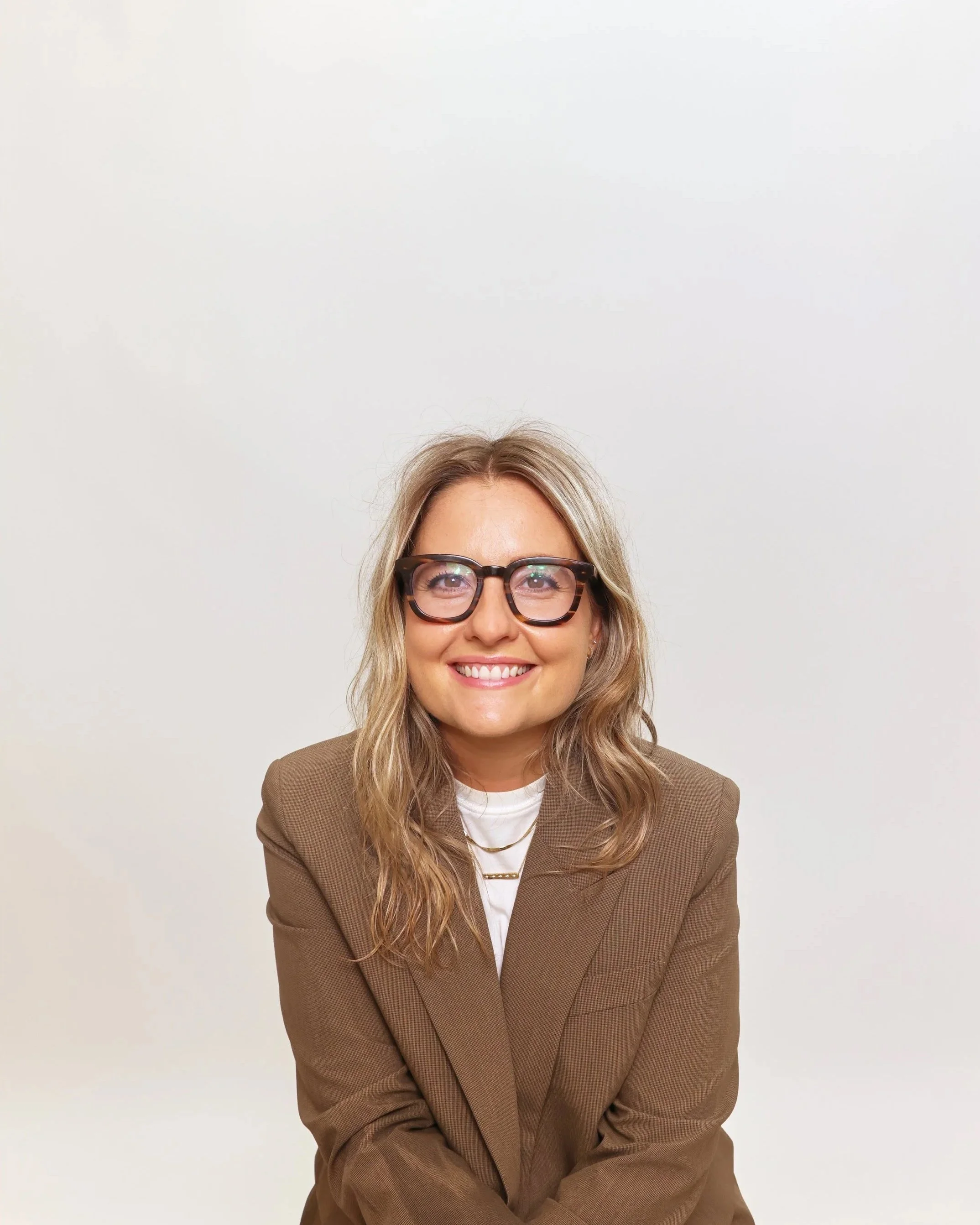 A woman with long wavy blonde hair, wearing glasses with thick tortoiseshell frames, smiling, dressed in a brown blazer over a white shirt, against a plain white background.