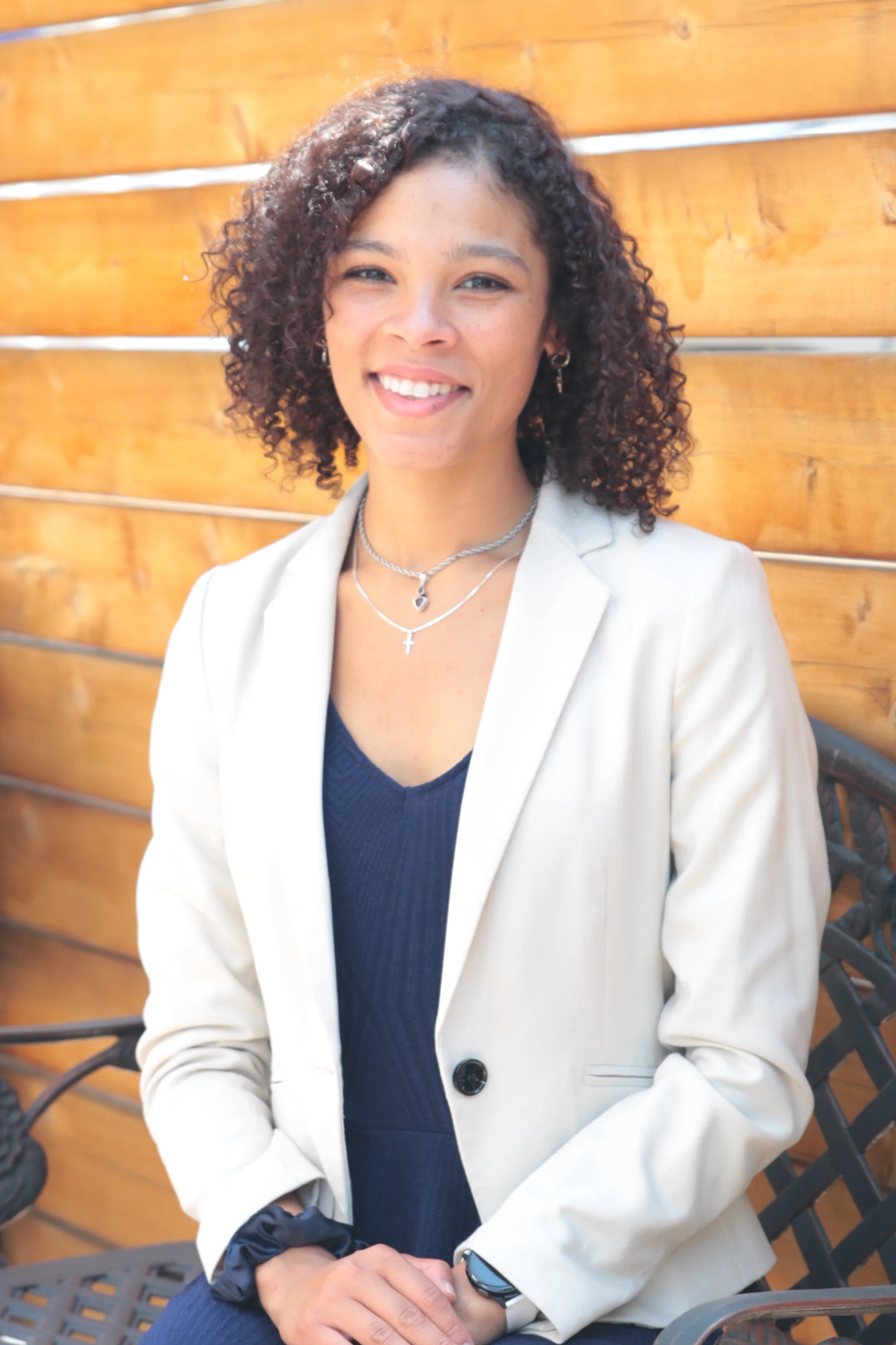 A smiling woman with curly hair, wearing earrings, layered necklaces, a beige blazer over a dark blue top, and sitting outdoors in front of a wooden fence.