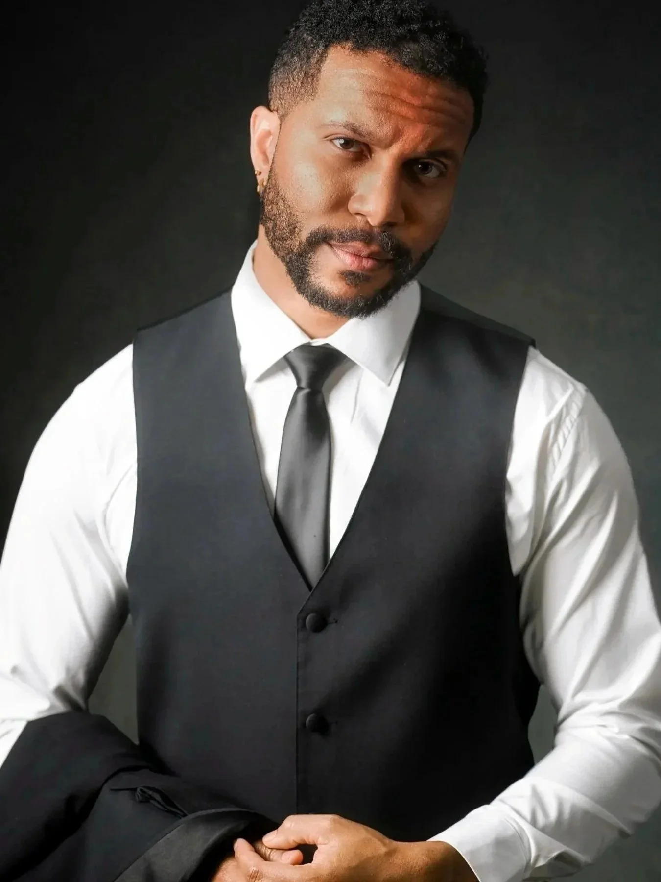 Portrait of a man in formal attire, wearing a white shirt, black vest, and black tie, with short curly hair and a beard, against a dark background.