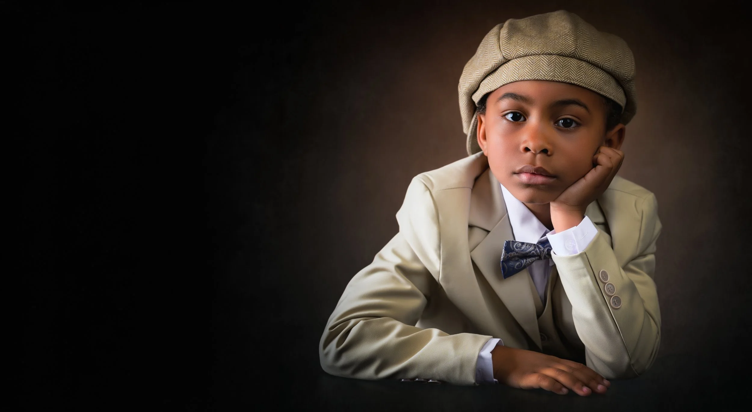 Young boy wearing a beige suit, white shirt, and bow tie, resting his chin on his hand with thoughtful expression, against a dark background.