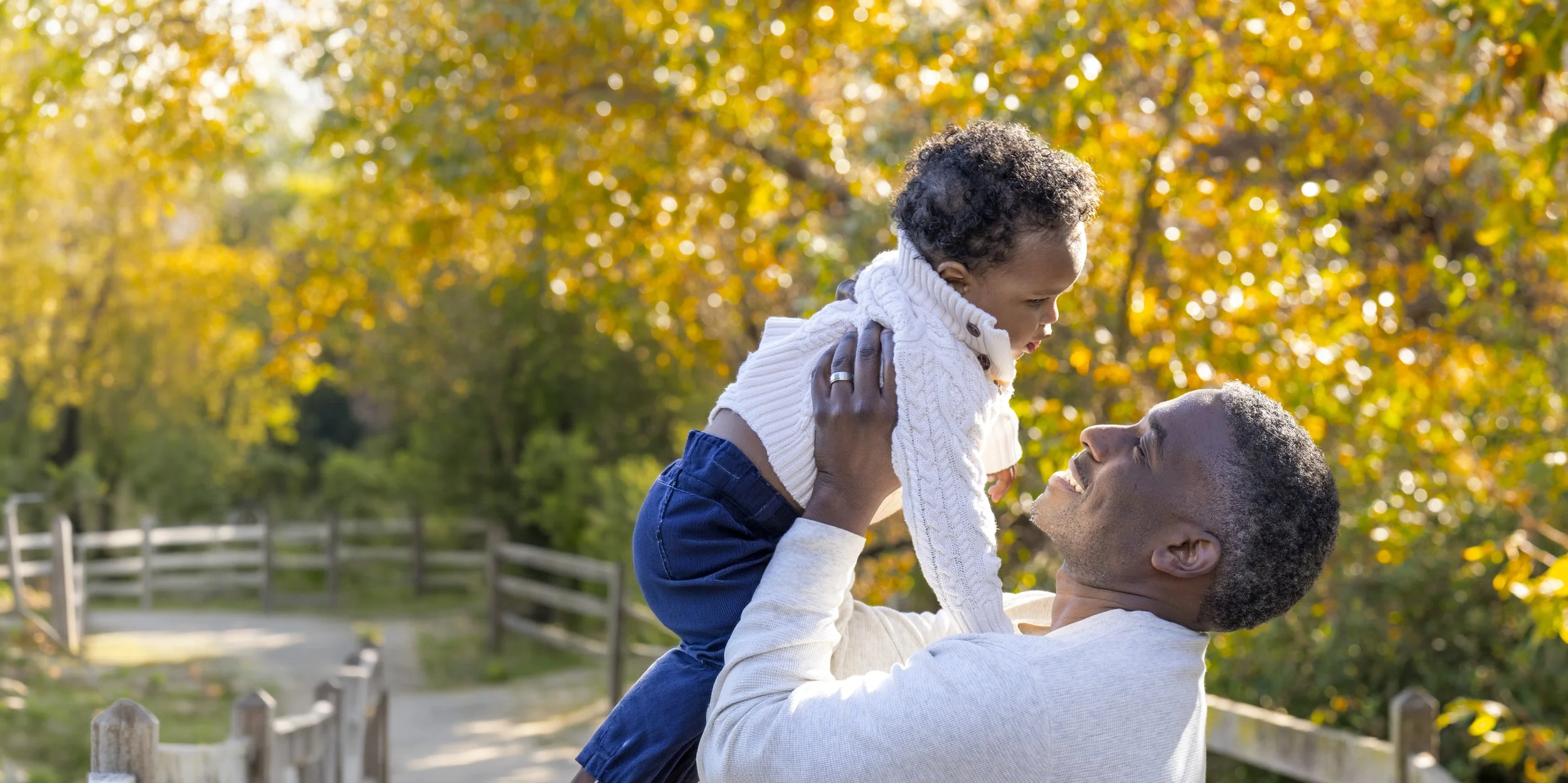 A man lifting a young child in a park during fall, with trees showing colorful autumn leaves in the background.