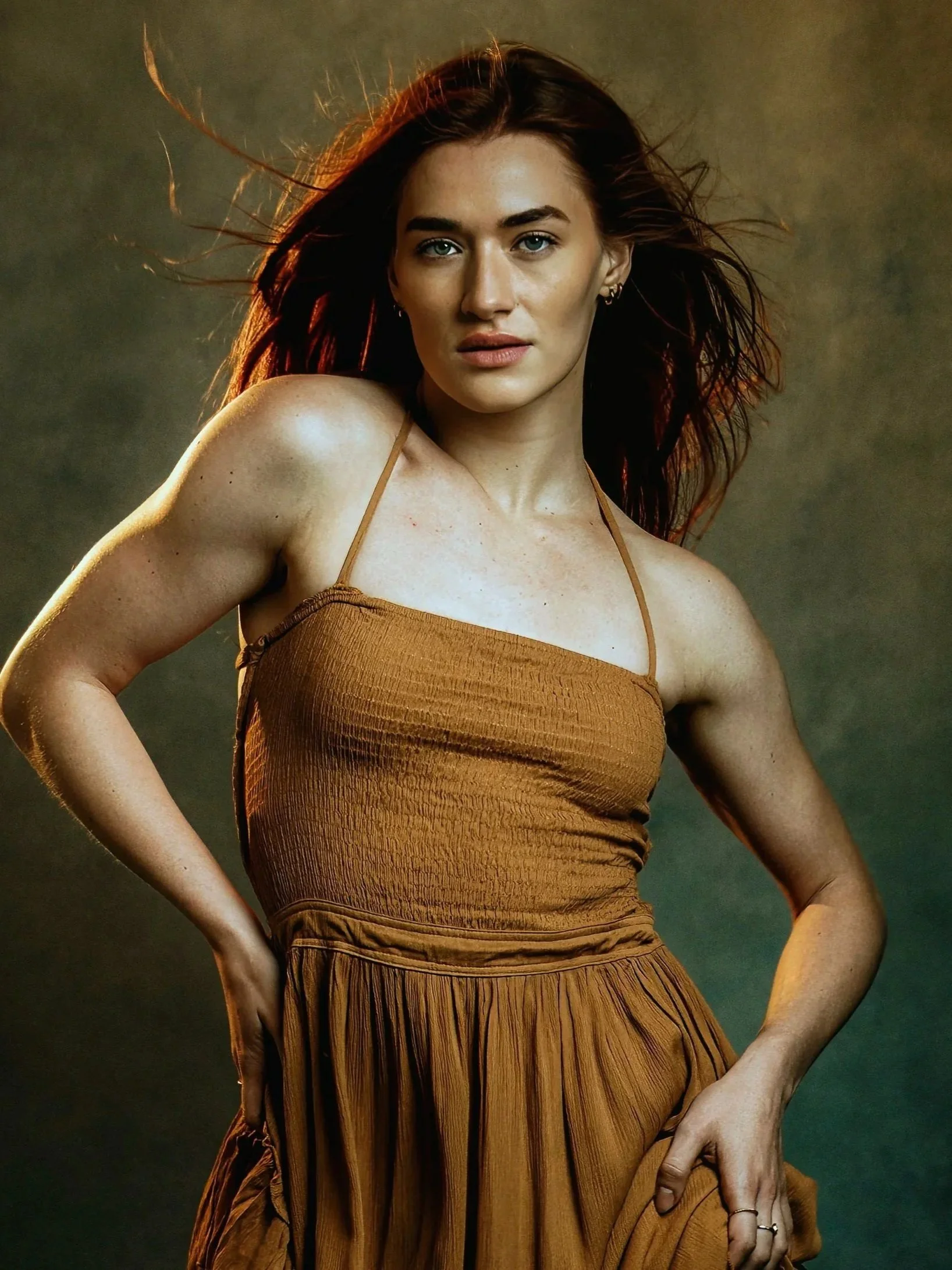 A woman with red hair flowing, wearing a brown dress with thin straps, posed with one hand on her hip against a dark background.