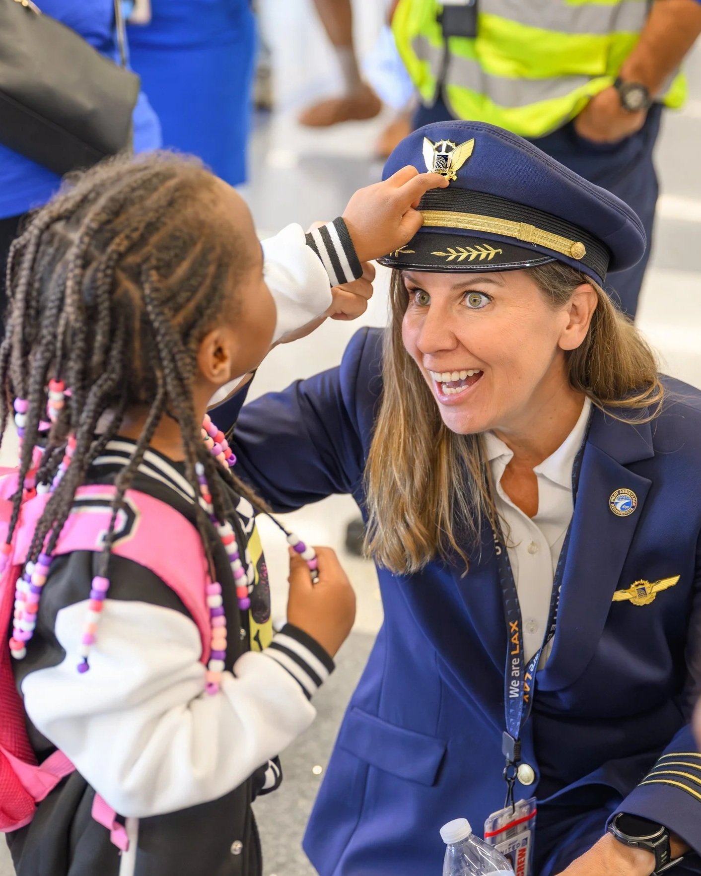 A young girl with braided hair and a pink backpack touches the brim of a female airline pilot's cap while the pilot smiles and looks at her, seated in an airport or similar setting.